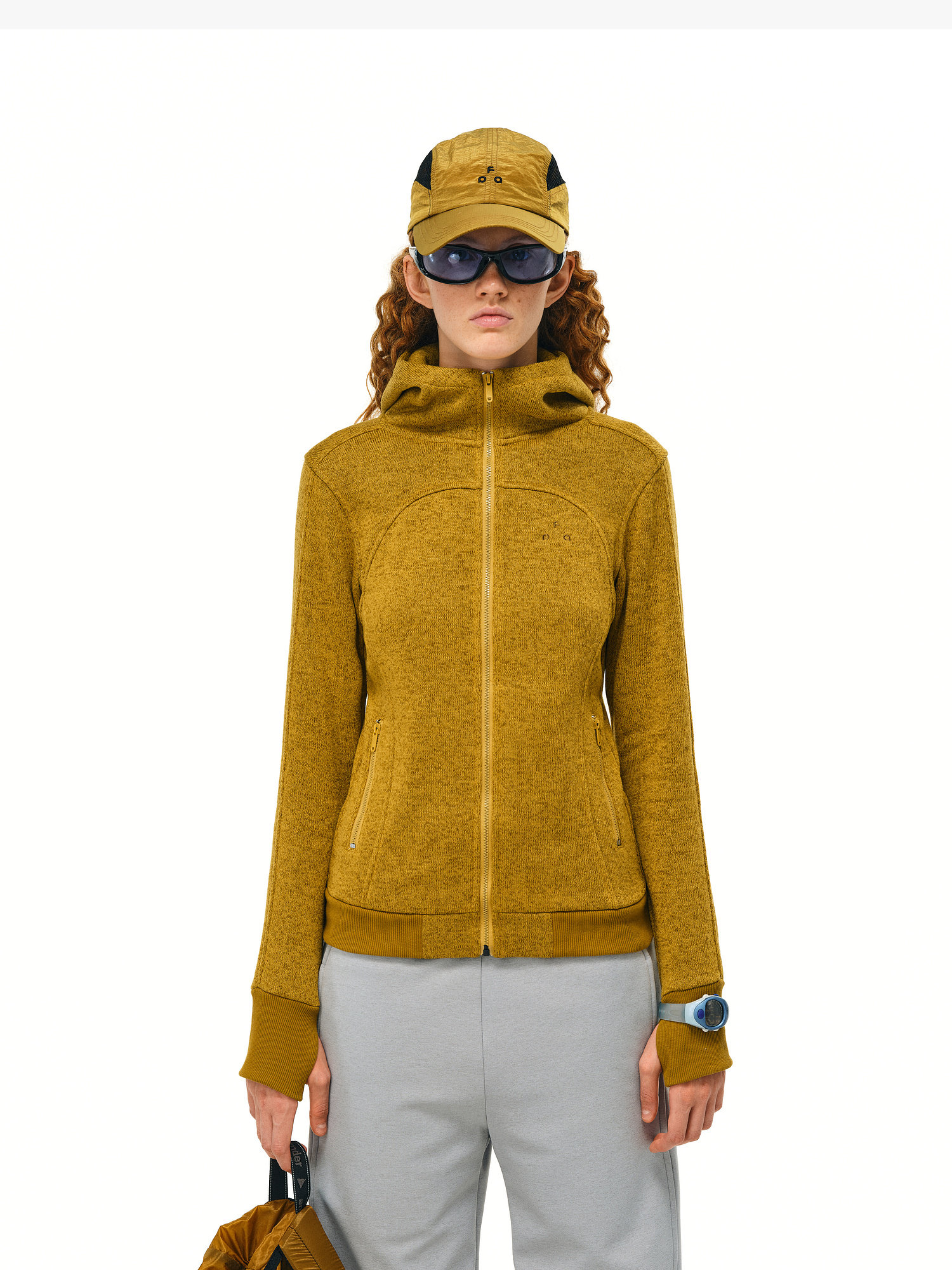 A person with curly hair wears an EastLyst Cropped Hooded Zip-Up Fleece Jacket in mustard yellow, a matching cap, sunglasses, and light gray pants. They hold a matching bag and wear a smartwatch, standing against a plain white background.
