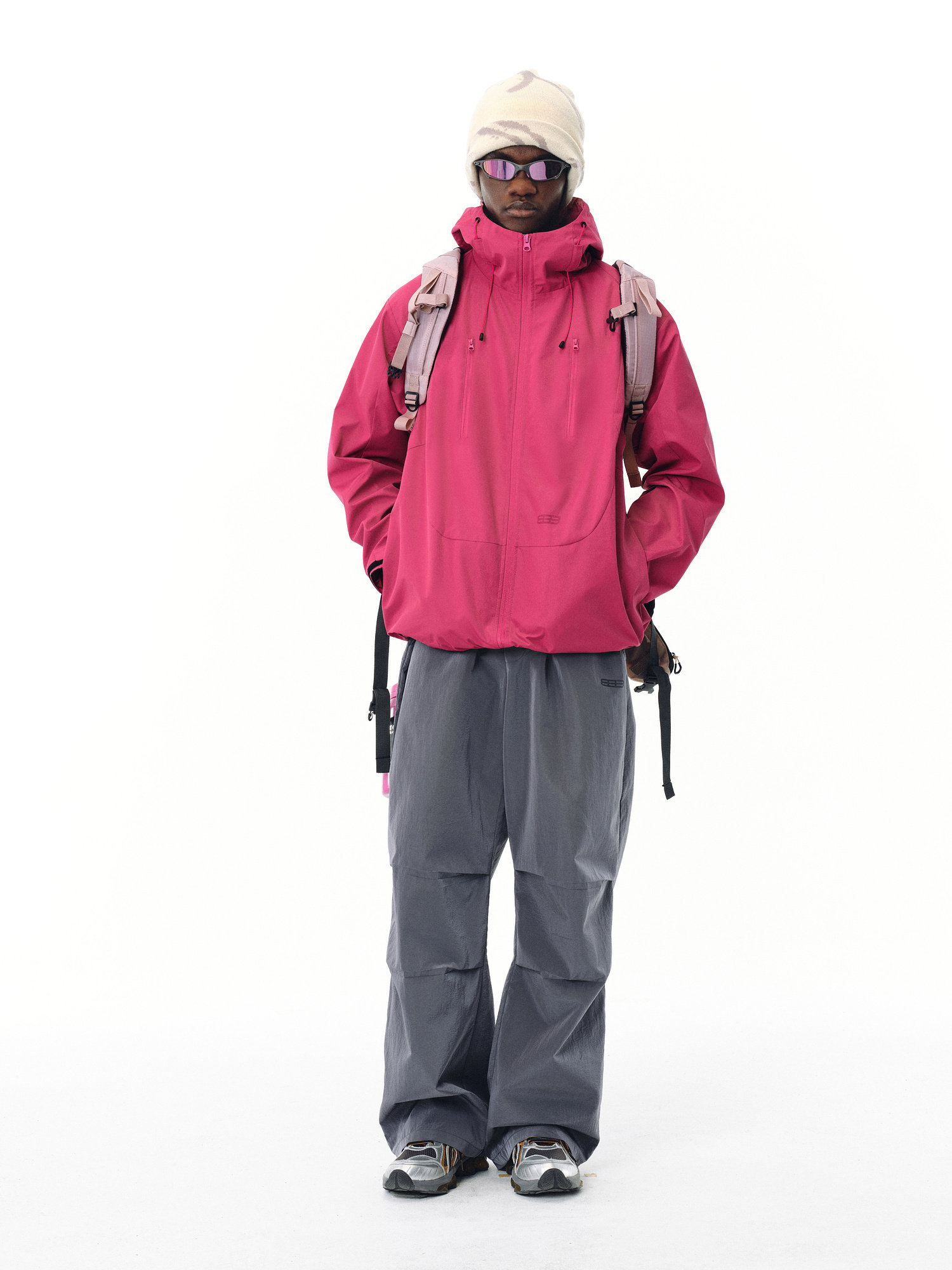 A person stands facing forward in the EastLyst Best All-Rounder Jacket (pink), gray baggy pants, gray sneakers, a white beanie, sunglasses, and a pink backpack against a plain white background.