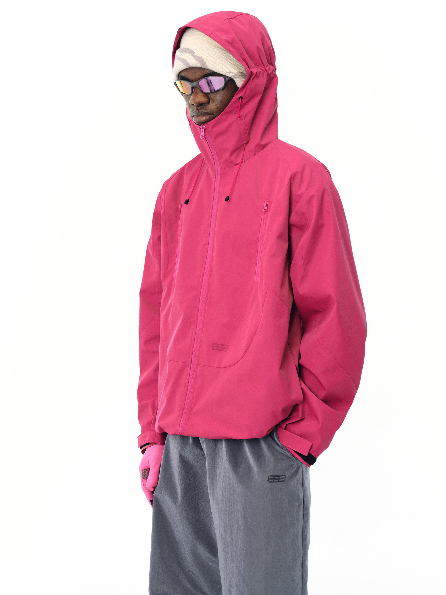 A person stands against a plain white background wearing the EastLyst Best All-Rounder Jacket in bright pink, paired with pink gloves, sunglasses, a beige hat, and loose gray pants with their hands in their pockets.