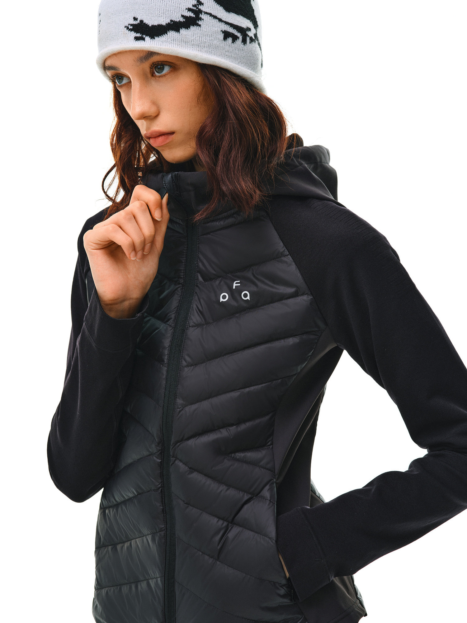 A young woman wears the EastLyst Quilted Hooded Down Jacket and a white beanie with a black pattern, standing against a white background with one hand on her collar, highlighting its lightweight warmth and style.