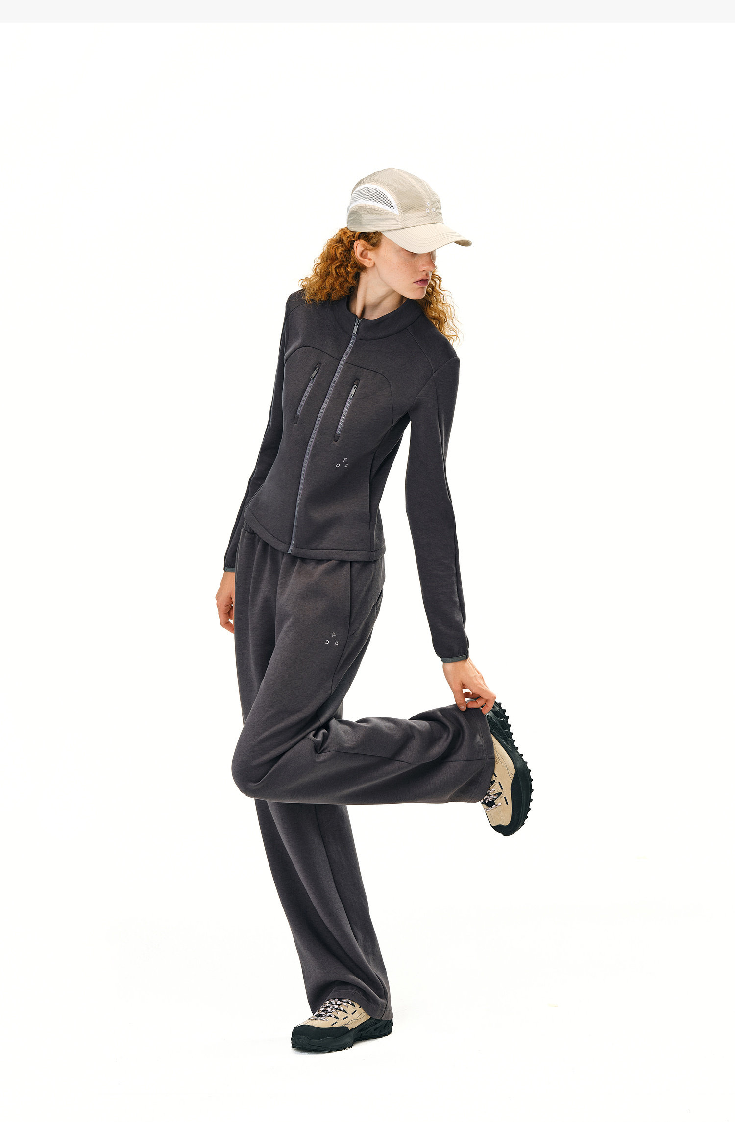 A curly-haired person wearing EastLyst Double-Layer Air-Knit Wide Leg Pants poses on one leg in a light cap and dark zip-up jacket, set against a white background.