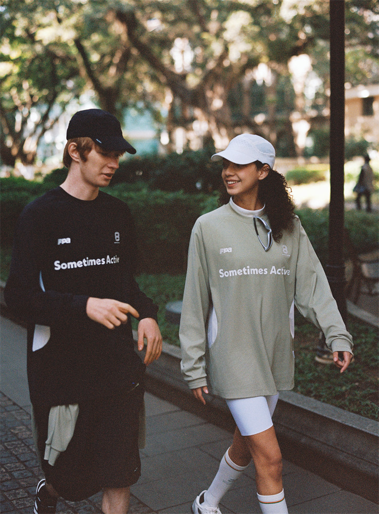 Two people stroll side by side in a park, smiling and talking amid trees. Both wear caps and EastLyst Quick-Dry Color Block Long Sleeve Tees printed with "Sometimes Active.