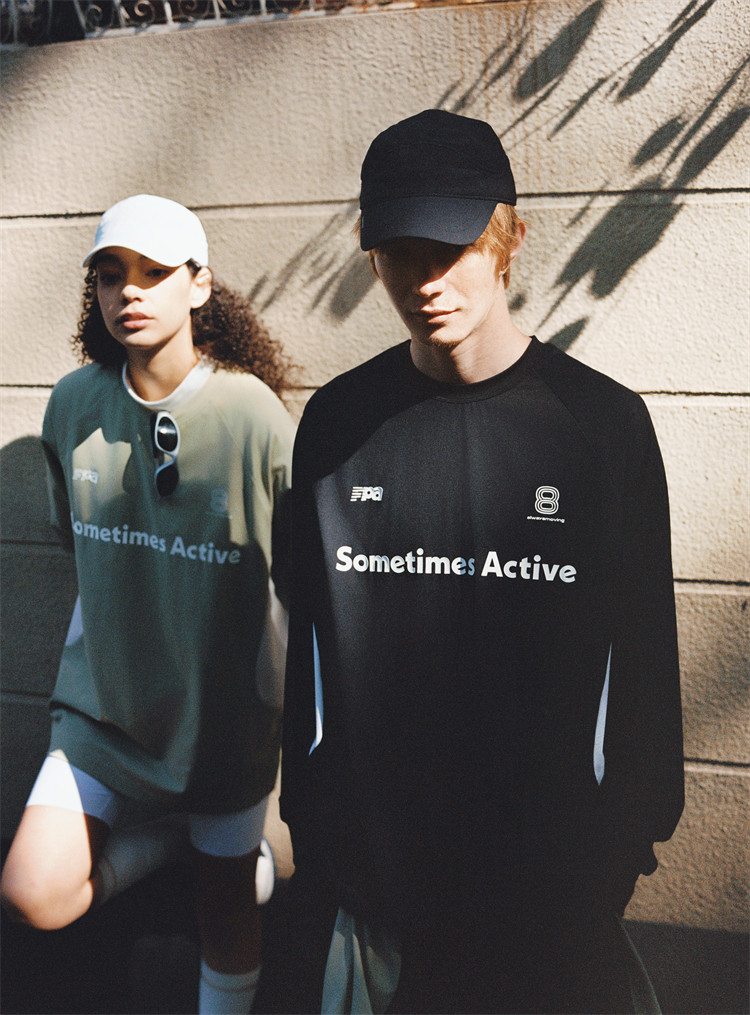 Two people in sporty outfits stand against a sunlit wall, each wearing an EastLyst Quick-Dry Color Block Long Sleeve Tee—one in green with a white cap, the other in black with a black cap.