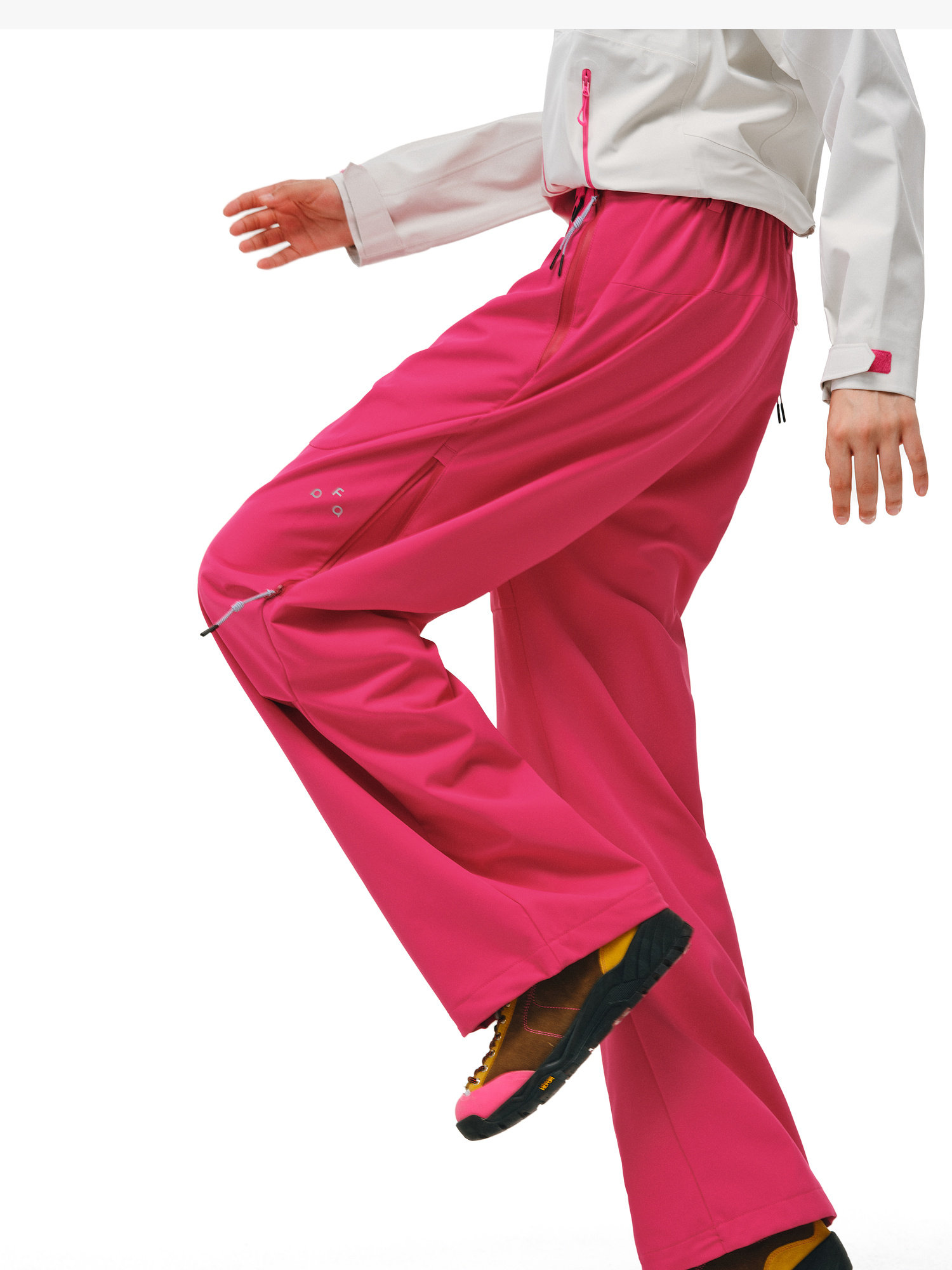 A person steps forward against a white background, showcasing EastLyst Softshell Trousers for Hiking in bright pink. They are styled with a white jacket and black and yellow shoes, with their head out of frame.