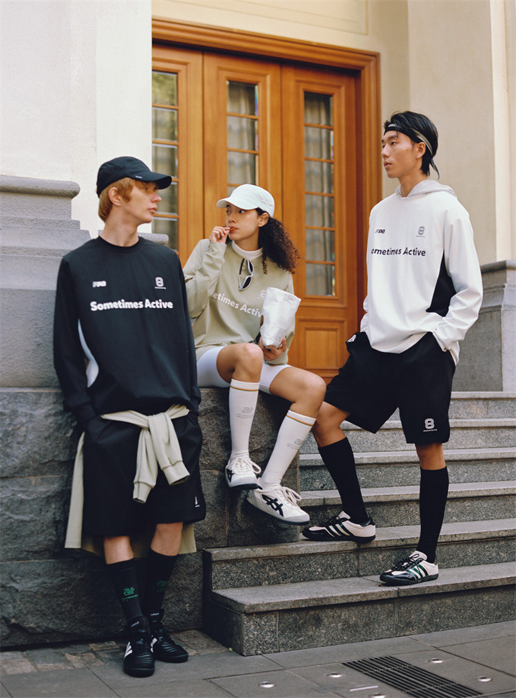 Three young people in EastLyst Quick-Dry Color Block Long Sleeve Tees pose outside a building with wooden doors. Two lean on a stone ledge while the third sits nearby, wearing sporty shorts and caps and eating from a bag.