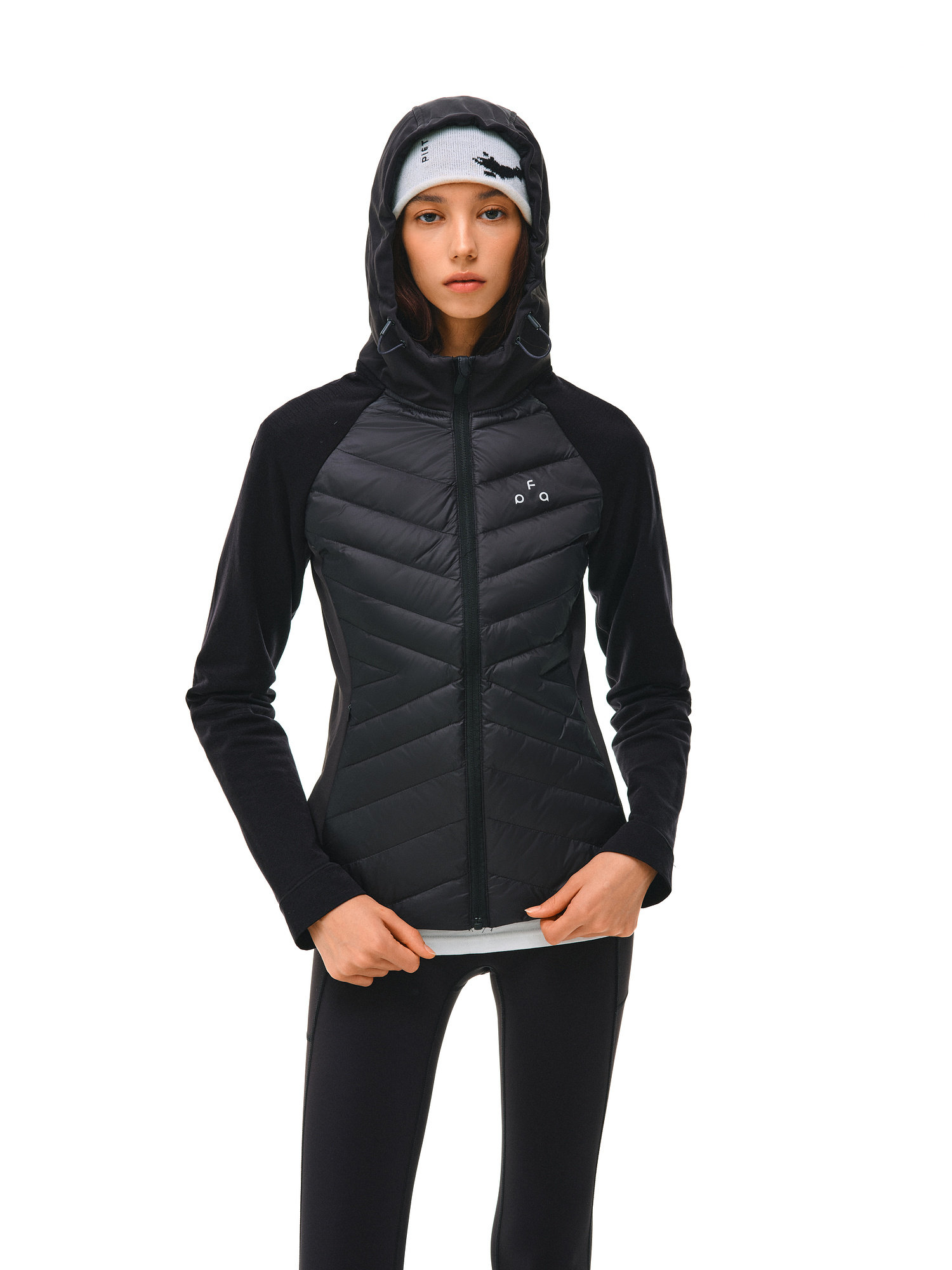 A person stands facing forward against a white background, hands near the zipper of an EastLyst Quilted Hooded Down Jacket, paired with black athletic pants and a light gray headband.