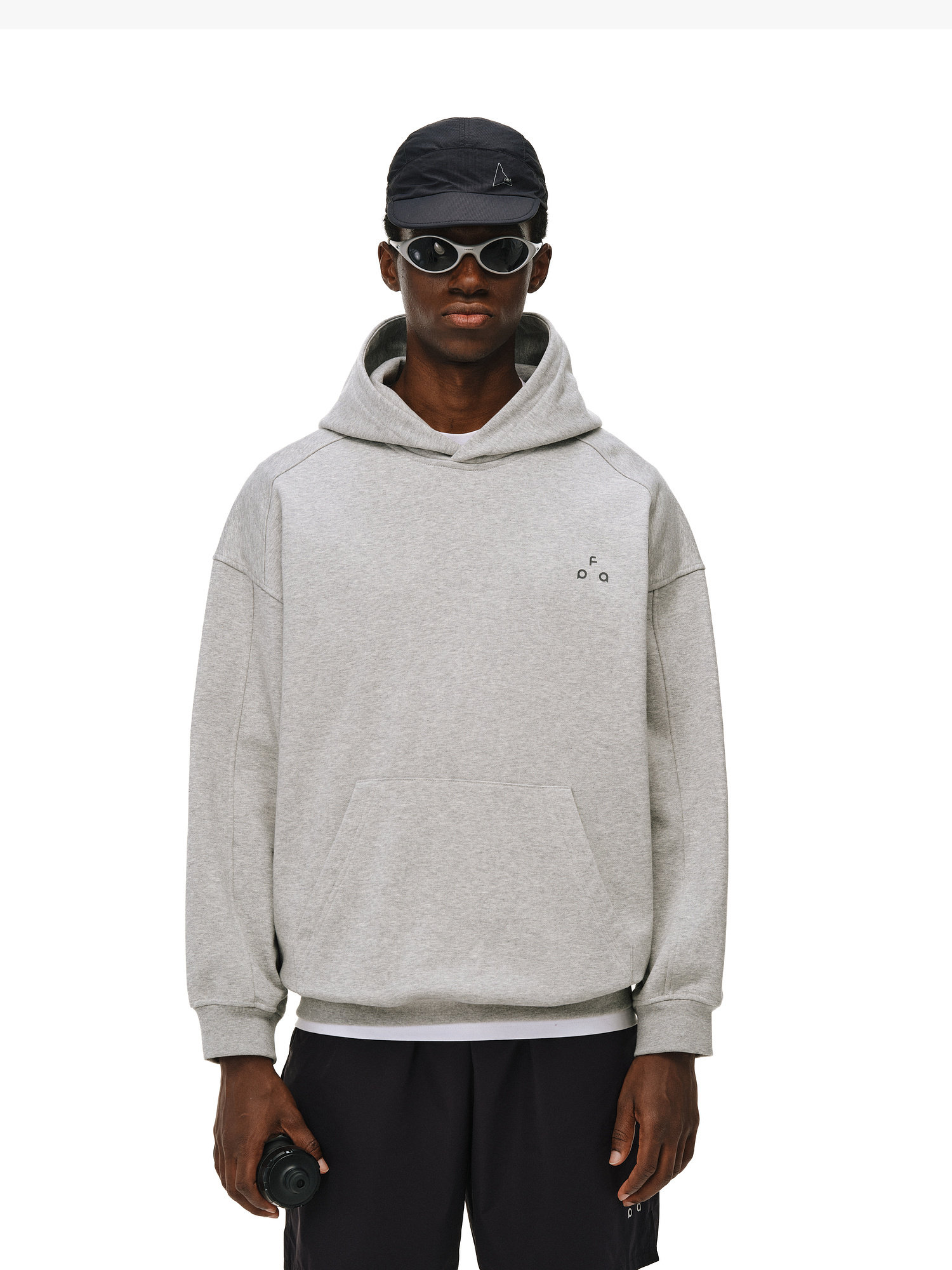 A person wearing an EastLyst Outdoor Print Hoodie, black sunglasses, and a black cap stands against a plain white background, holding a small black object in their right hand.