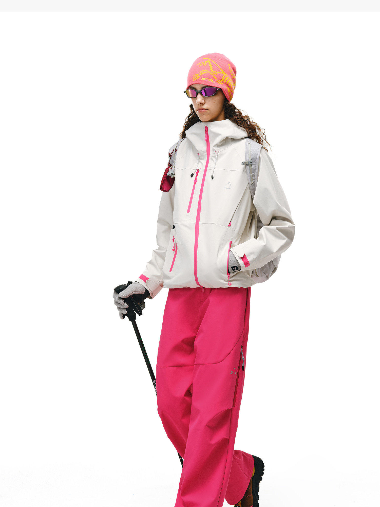 A person wears a white jacket, EastLyst Softshell Trousers for Hiking in bright pink, a pink beanie, and sunglasses, holding ski poles against a plain white background.