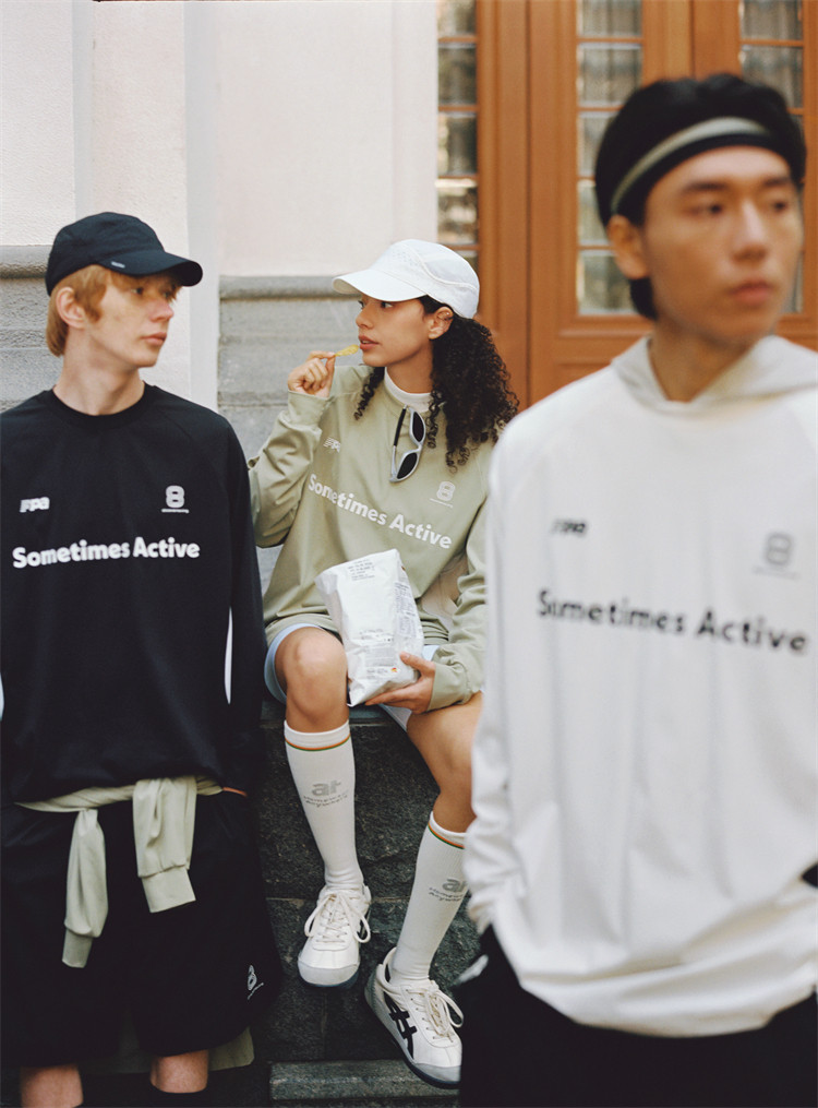 Three young adults in sporty, casual outfits by EastLyst stand and sit outdoors. Two wear “Sometimes Active” sweatshirts, while the third adds lightweight comfort to the scene in an EastLyst Quick-Dry Color Block Long Sleeve Tee.