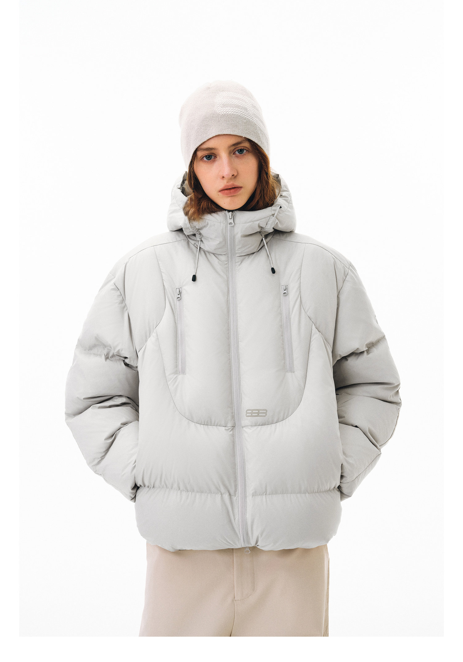 A person models the EastLyst Fashionable 3D short down jacket in light gray with a hood, paired with a beige beanie and pants, standing against a plain white background and looking directly at the camera with hands in pockets.