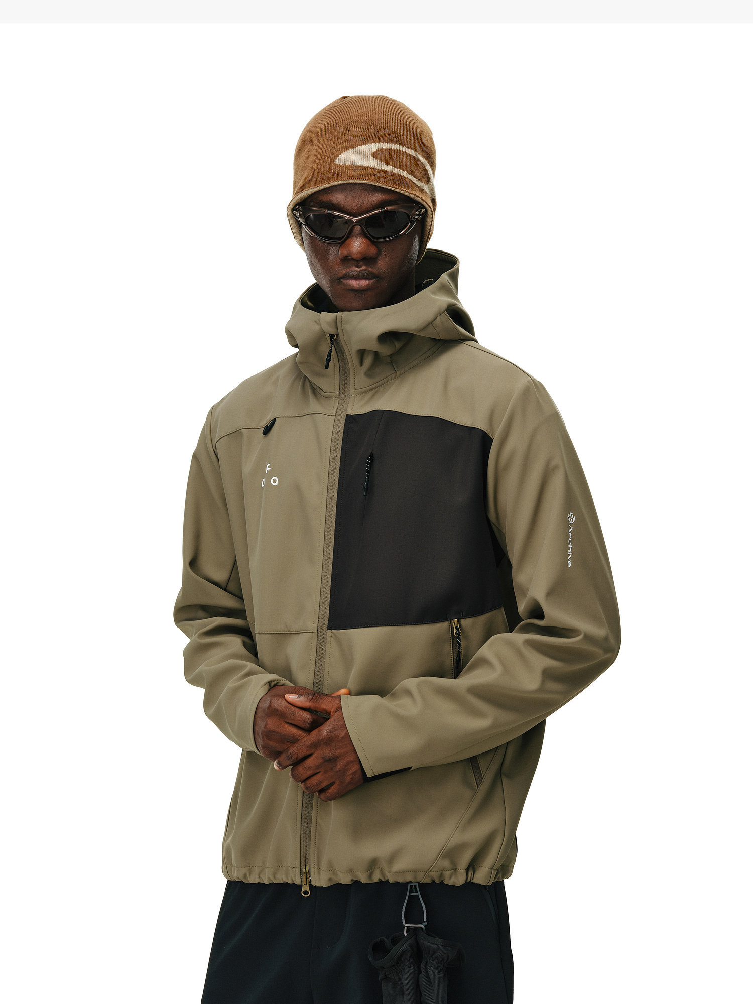 A person wearing a brown beanie, black sunglasses, and an EastLyst Color-Block Water-Resistant Softshell Jacket with a black chest pocket stands against a white background with hands clasped in front.