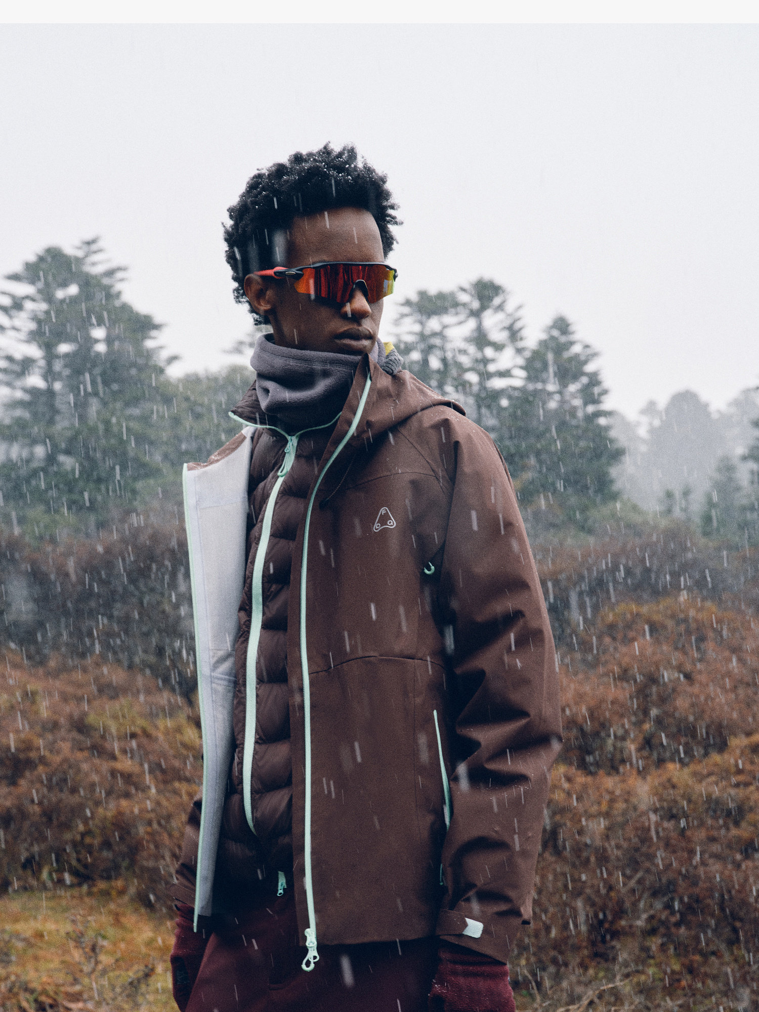 Outdoors in falling snow, a person wears the EastLyst 3-in-1 Color-Block Storm Jacket with layered clothing, gloves, and reflective sunglasses—staying warm and stylish for any urban commute amid misty trees and bushes.