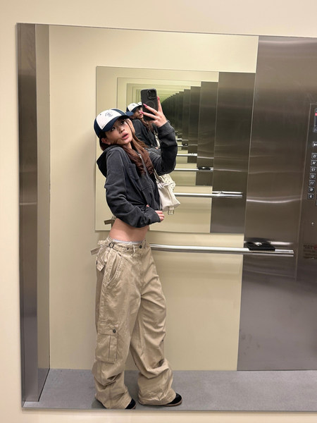 Model wearing a casual layered streetwear outfit with a dark jacket, baseball cap, and beige cargo pants.