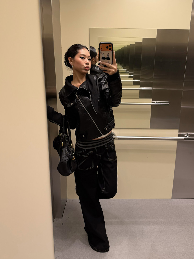 Person wearing a black leather jacket, black cargo pants, and carrying a black shoulder bag in an elevator mirror selfie.