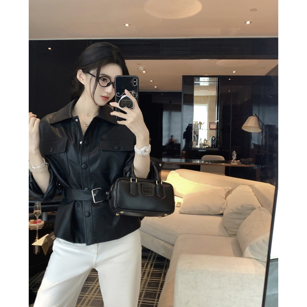 Woman taking mirror selfie wearing black leather shirt with belt and white pants, holding black handbag
