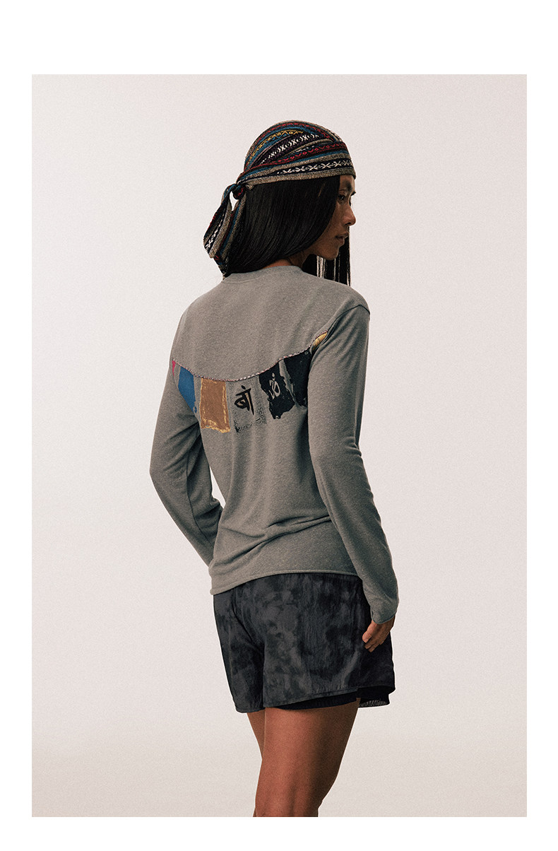 A woman faces away from the camera wearing an EastLyst Vintage vintage T-shirt with colorful prayer flag patches on the back, dark shorts, and a patterned headscarf.