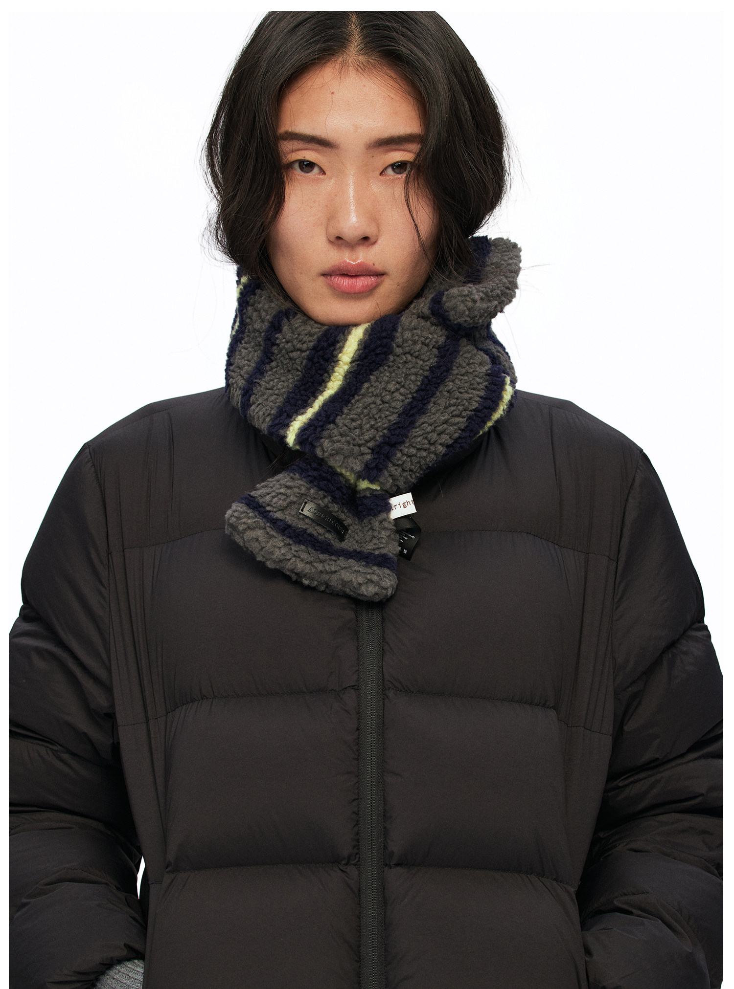 A person with long dark hair wears a black puffer jacket and an EastLyst Striped Fleece Scarf, featuring navy and yellow stripes, standing against a plain white background.