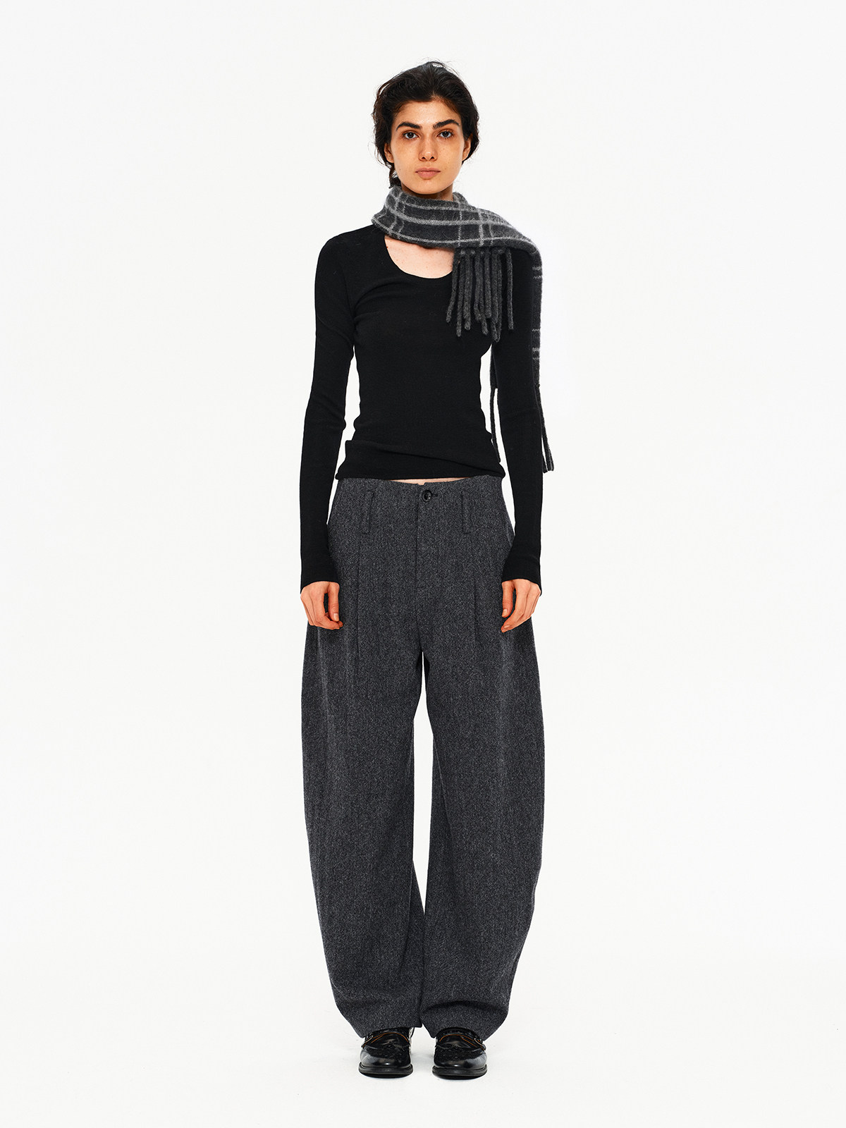 A person stands facing forward against a plain white background, wearing a black cutout long-sleeve top, wide gray trousers, black shoes, and an EastLyst Karl alpaca wool classic scarf draped over their shoulders.