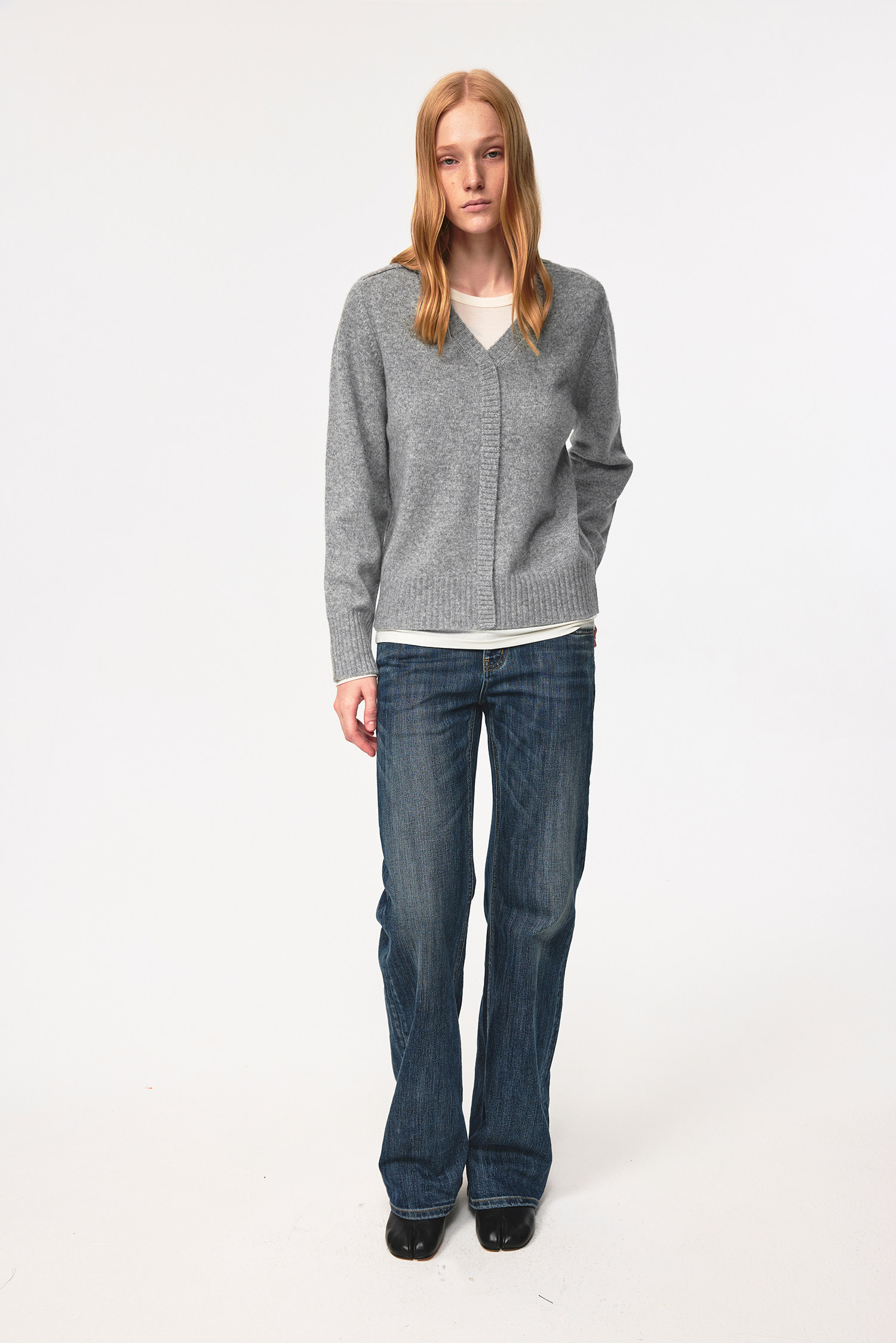 A person with long blond hair stands facing forward, wearing a grey V-neck sweater over a white shirt, EastLyst Slub Textured Casual Pants in vintage indigo wash, and black shoes against a plain white background.