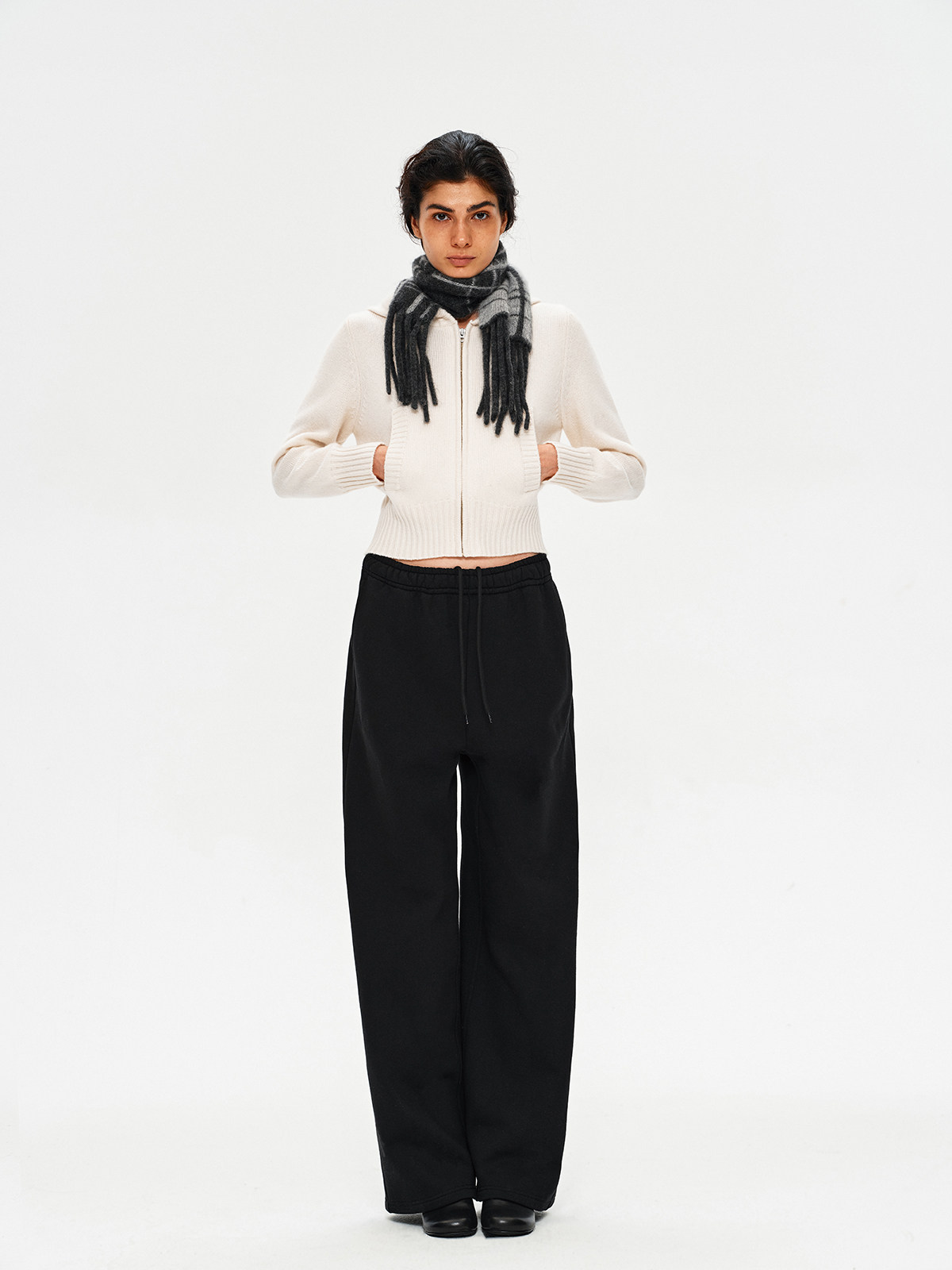 A person stands against a plain white background, hands in pockets, wearing a cream zip-up sweater, loose black trousers, black shoes, and the EastLyst Karl alpaca wool classic scarf.