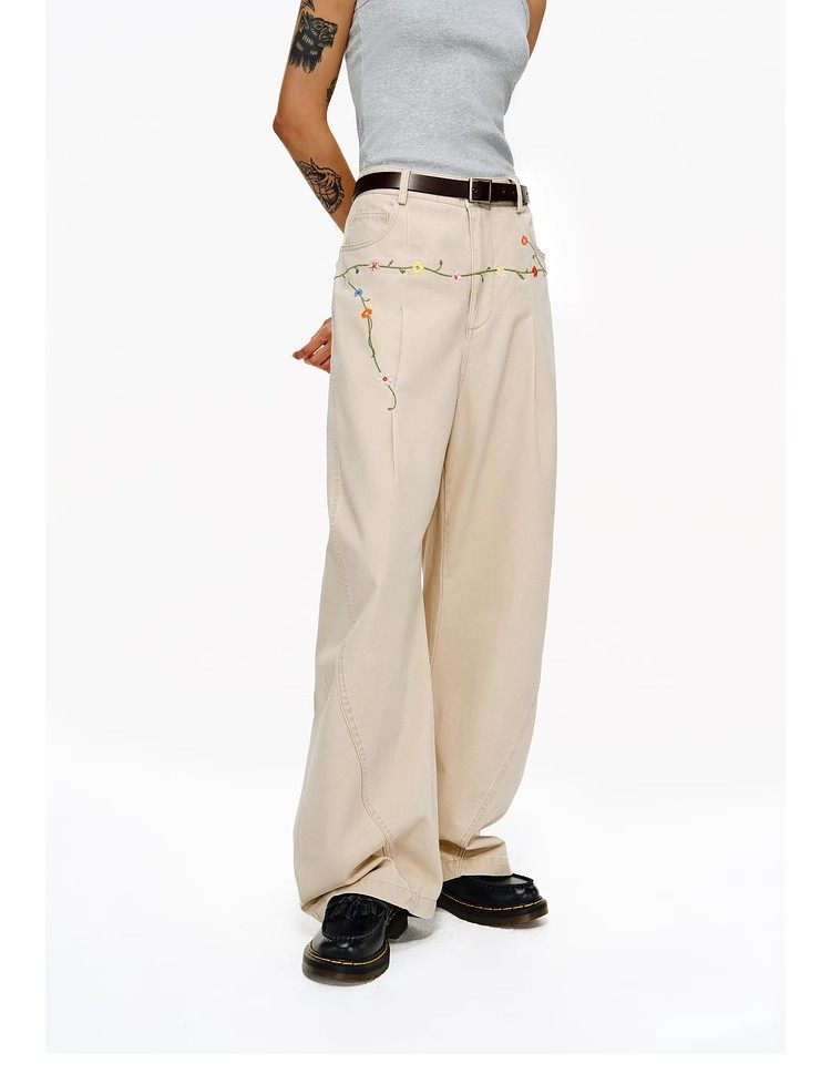 A person stands against a plain white background wearing EastLyst Floral Embroidery Cargo Pants, a gray sleeveless top, black shoes, and a brown belt.