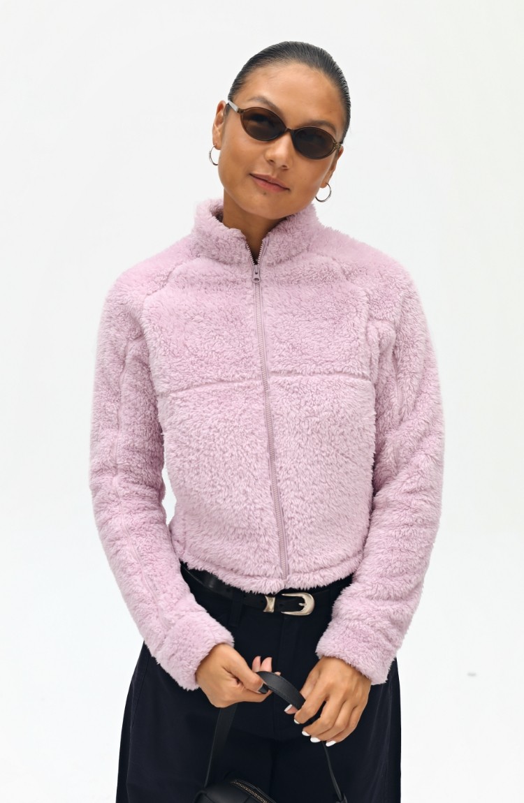 A person wears an EastLyst Pink Short Faux Shearling Jacket, black sunglasses, and hoop earrings, standing against a plain white background while holding a black strap with both hands.