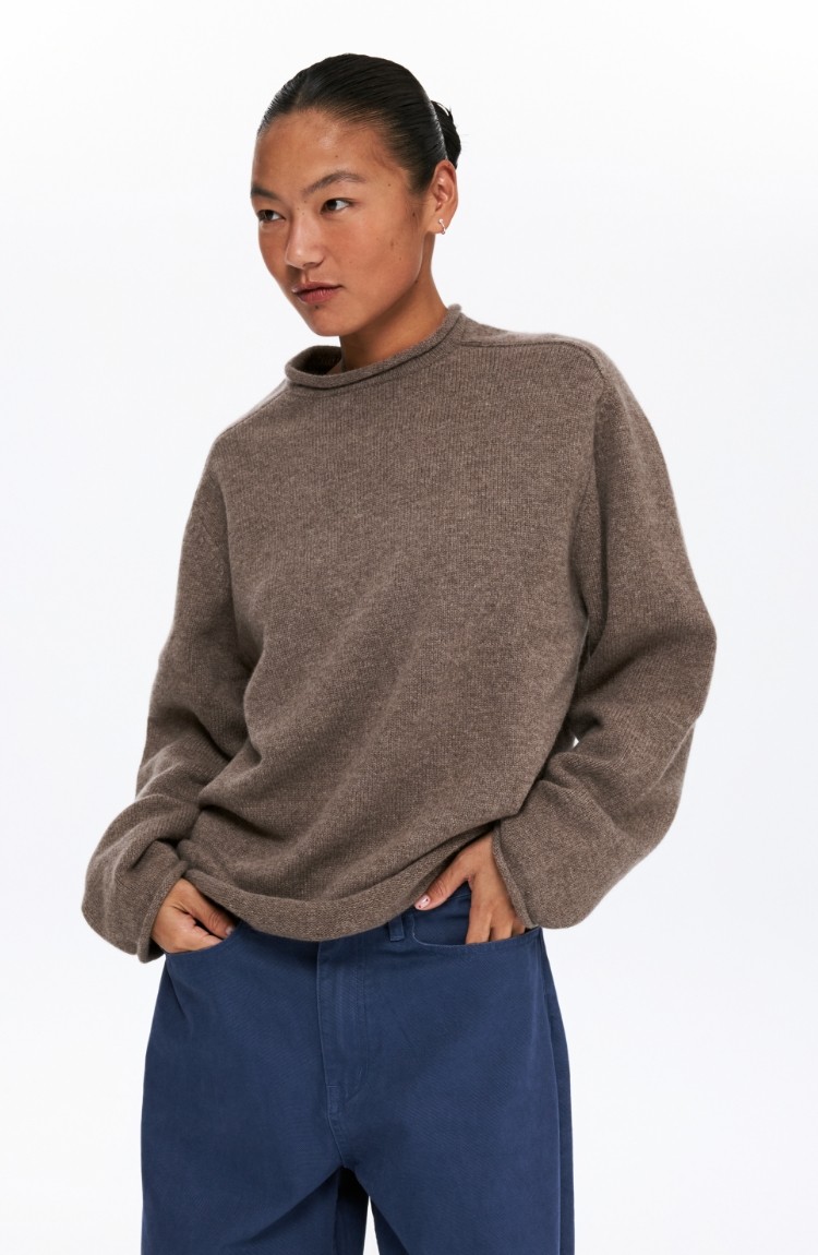 A person with dark hair tied back models the EastLyst Lounge Knit Sweater in brown and blue pants, standing casually with one hand in their pocket against a plain white background.