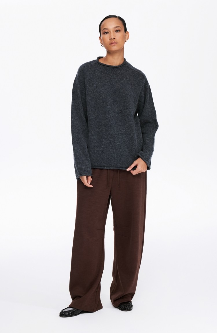 A person stands facing forward, wearing the EastLyst Lounge Knit Sweater in dark gray (100% wool), wide-legged brown pants, and black shoes. Their hair is tied back, hands at their sides, against a plain white background.