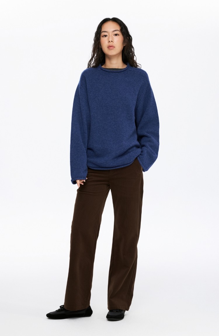 A person with long wavy hair stands facing forward, wearing the EastLyst Lounge Knit Sweater in blue, brown wide-leg pants, and black shoes against a plain white background.