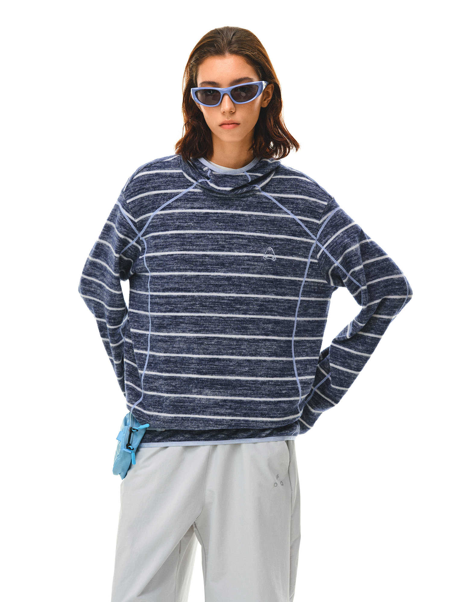 A person wears blue-tinted sunglasses, light gray pants, and the EastLyst Textured Stripe Knit Hoodie—a navy and white relaxed-fit, long-sleeve top—while standing with hands in pockets against a white background.