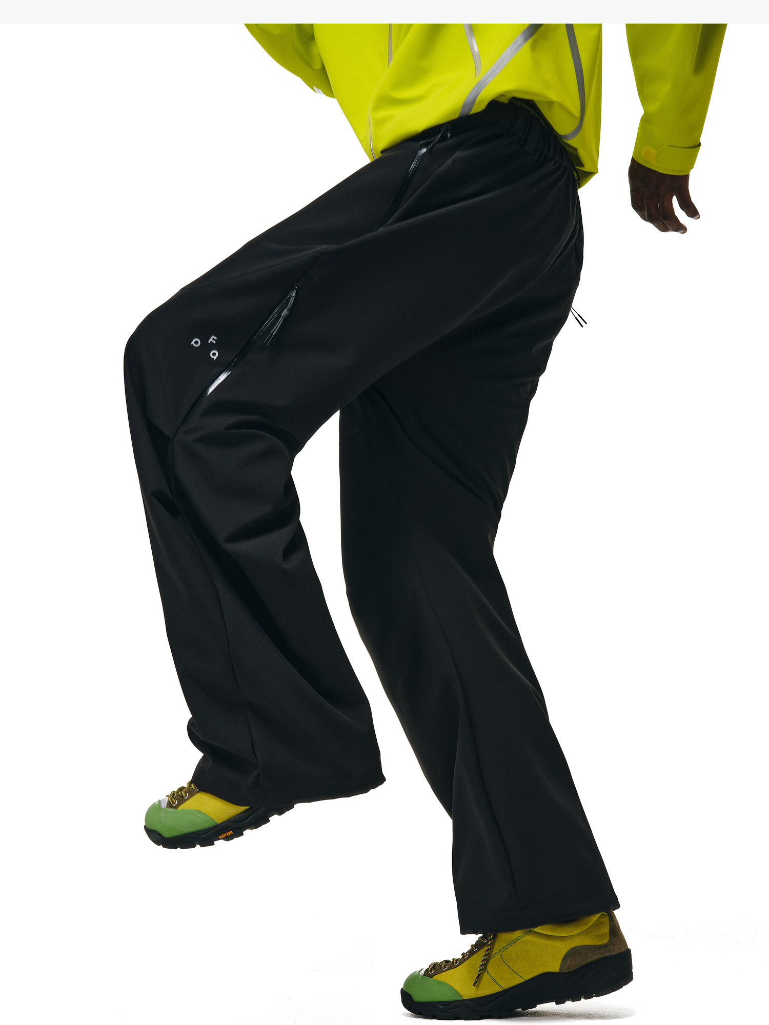 A person wearing EastLyst Softshell Joggers and green-yellow sneakers poses with one leg raised, styled in a neon yellow moisture-wicking, waterproof polyester jacket; the background is white.