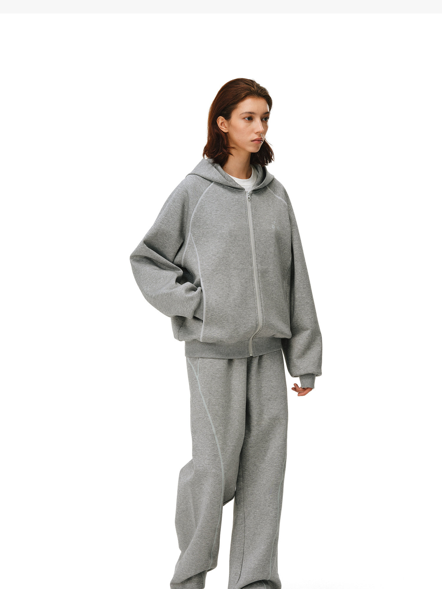 A woman with shoulder-length brown hair models the EastLyst Constant Warmth Zip Hoodie, an oversized light gray zip-up made from premium fabric, paired with matching wide-leg sweatpants against a plain white background.