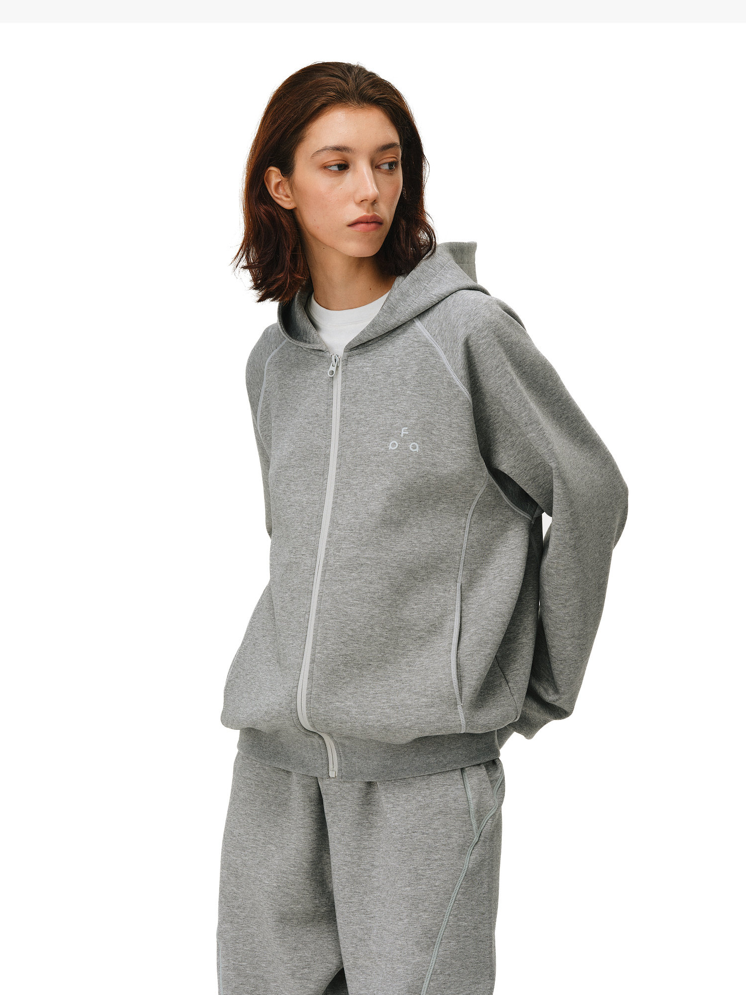 A person with shoulder-length brown hair models the EastLyst Constant Warmth Zip Hoodie and matching sweatpants in grey, crafted from high-end fabric, standing against a white background and looking to the side with a neutral expression.