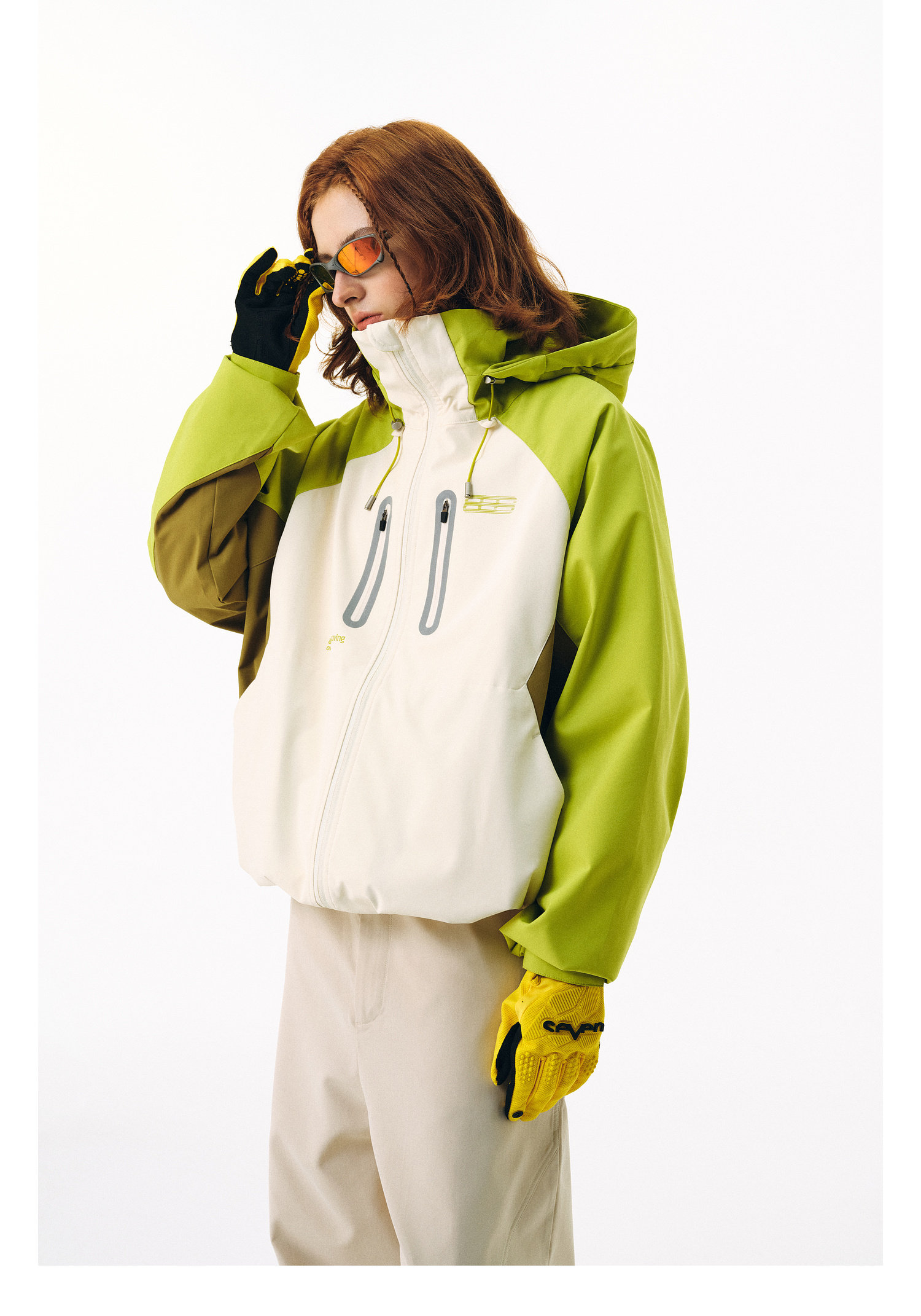 A person wears the EastLyst Versatile Explorer 3-in-1 Jacket—green and off-white, waterproof, with a hood—paired with beige pants, yellow gloves, and orange-tinted sunglasses against a plain white background.