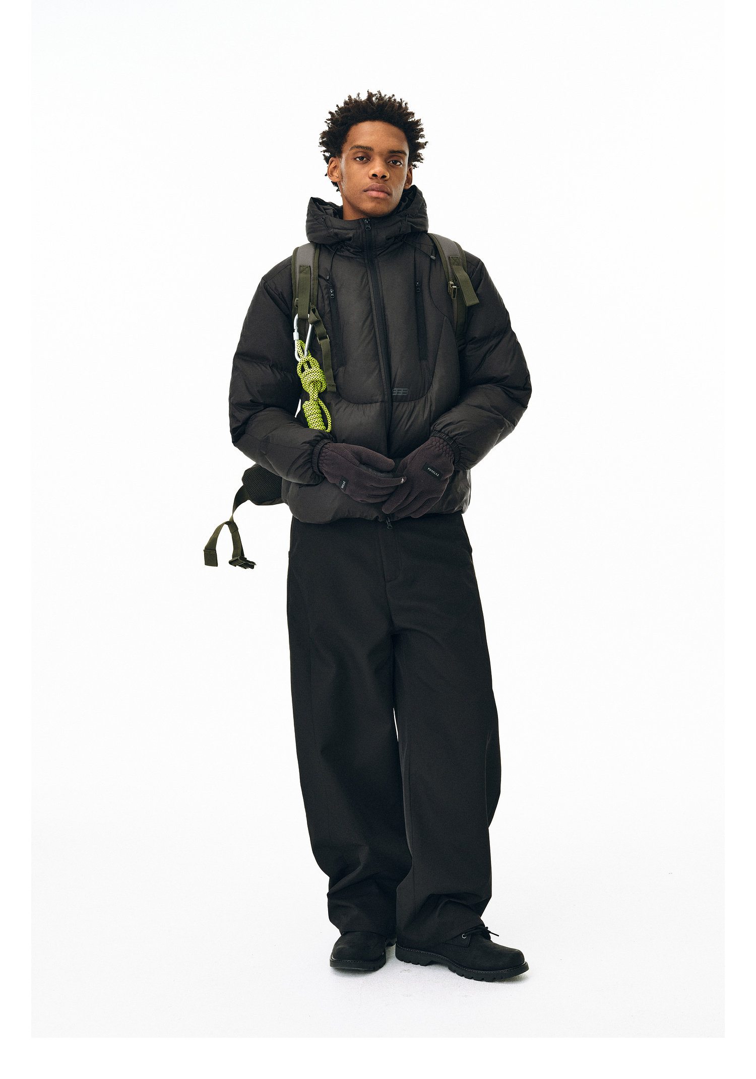 A young man stands against a white background wearing the EastLyst Fashionable 3D short down jacket, wide black pants, black gloves, shoes, and a green backpack with straps, looking slightly to the side.
