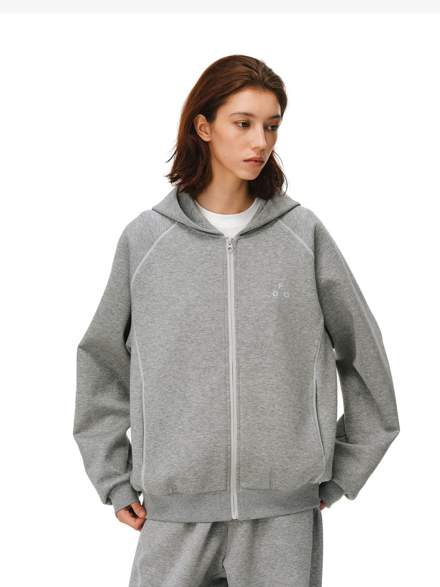A person with short brown hair models the EastLyst Constant Warmth Zip Hoodie and matching sweatpants in light gray, crafted from premium fabric, standing against a white background with hands in pockets, glancing slightly to the side.