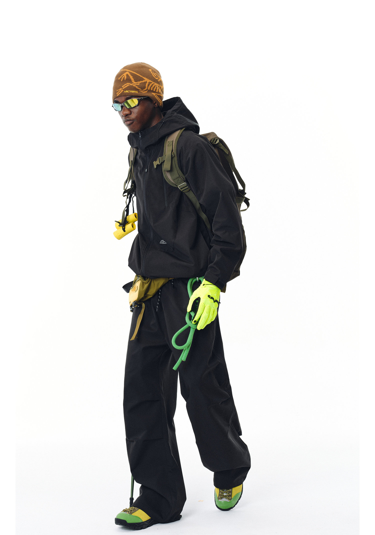 A person wears EastLyst’s Best All-Rounder Jacket (black, weather-resistant) with matching pants, green gloves and shoes, a brown patterned beanie, sunglasses, and carries green climbing gear plus a green backpack on a white background.