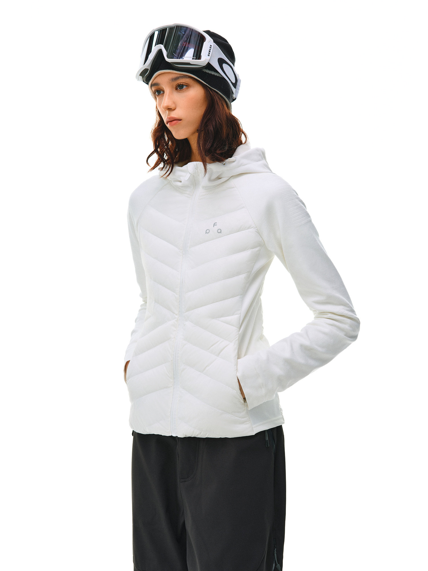 A woman models the EastLyst Quilted Hooded Down Jacket in white with black ski goggles and pants, hands in pockets, against a white background—highlighting lightweight warmth and contemporary style.