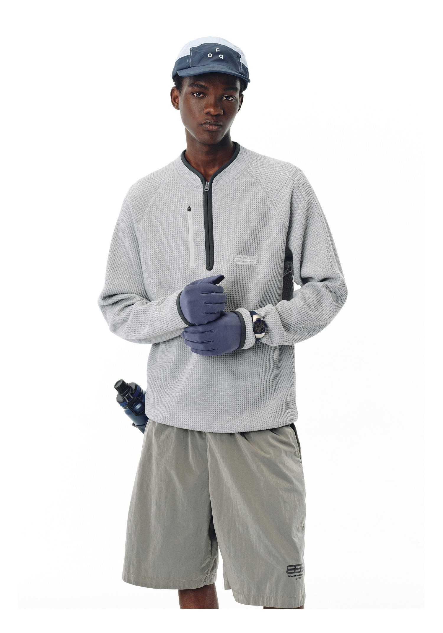 A man stands against a white background wearing an EastLyst Heavyweight Magnetic Waffle Knit Pullover Hoodie in light gray, gray shorts, a blue cap with cartoon eyes, blue gloves, a watch, and has a water bottle clipped to his shorts.
