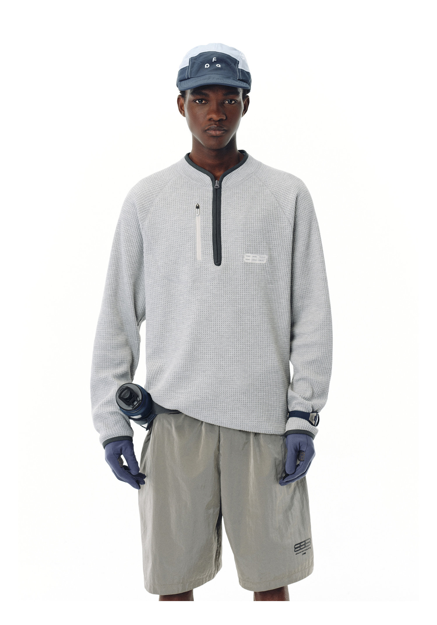 A person wears the EastLyst Heavyweight Magnetic Waffle Knit Pullover Hoodie, taupe shorts, blue gloves, a blue-gray cap, and holds a water bottle while standing against a plain white background.
