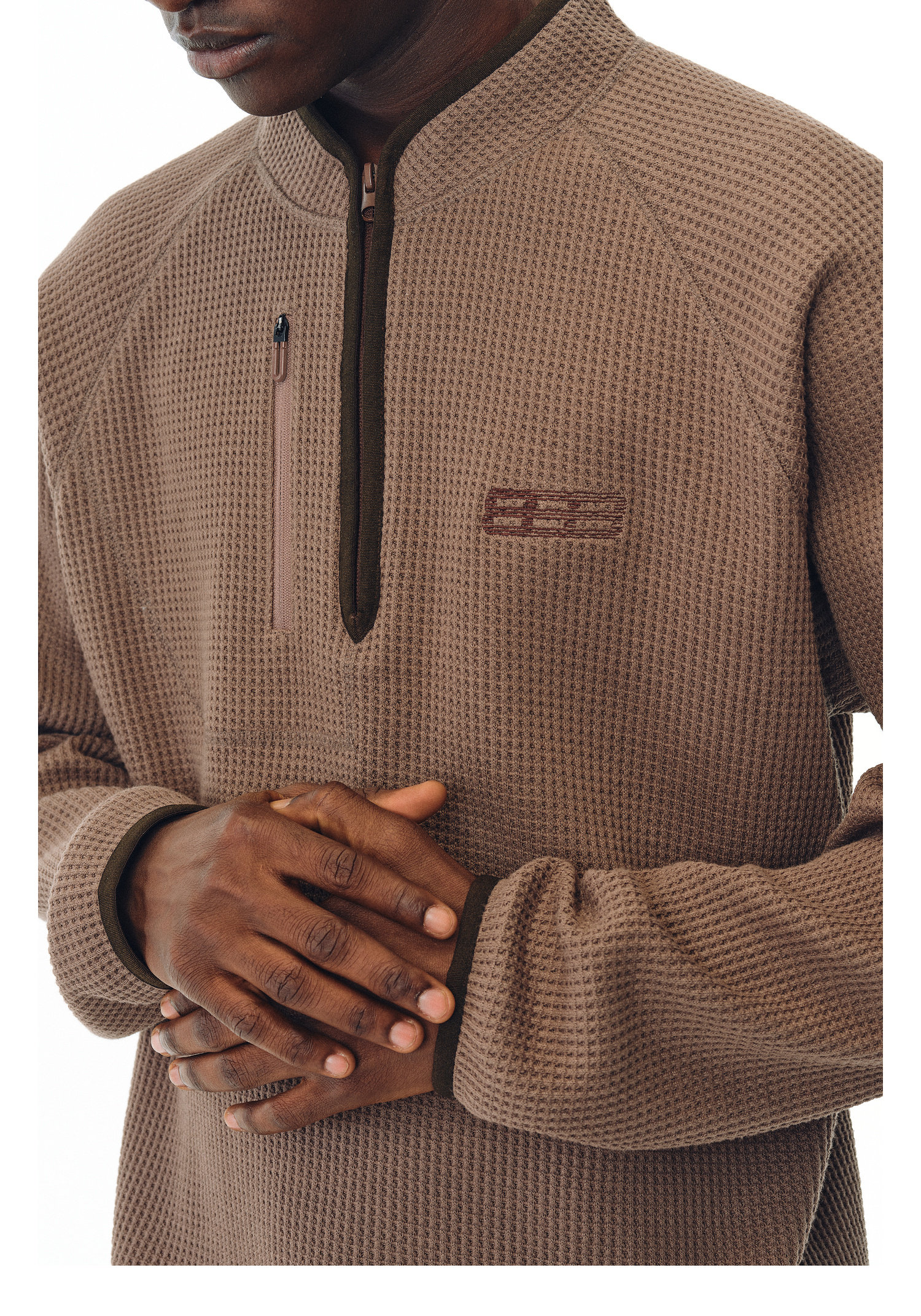 A person wearing the EastLyst Heavyweight Magnetic Waffle Knit Pullover Hoodie, featuring a textured brown fabric and zippered chest pocket, with their hands clasped. The image is cropped to show only the torso and hands.