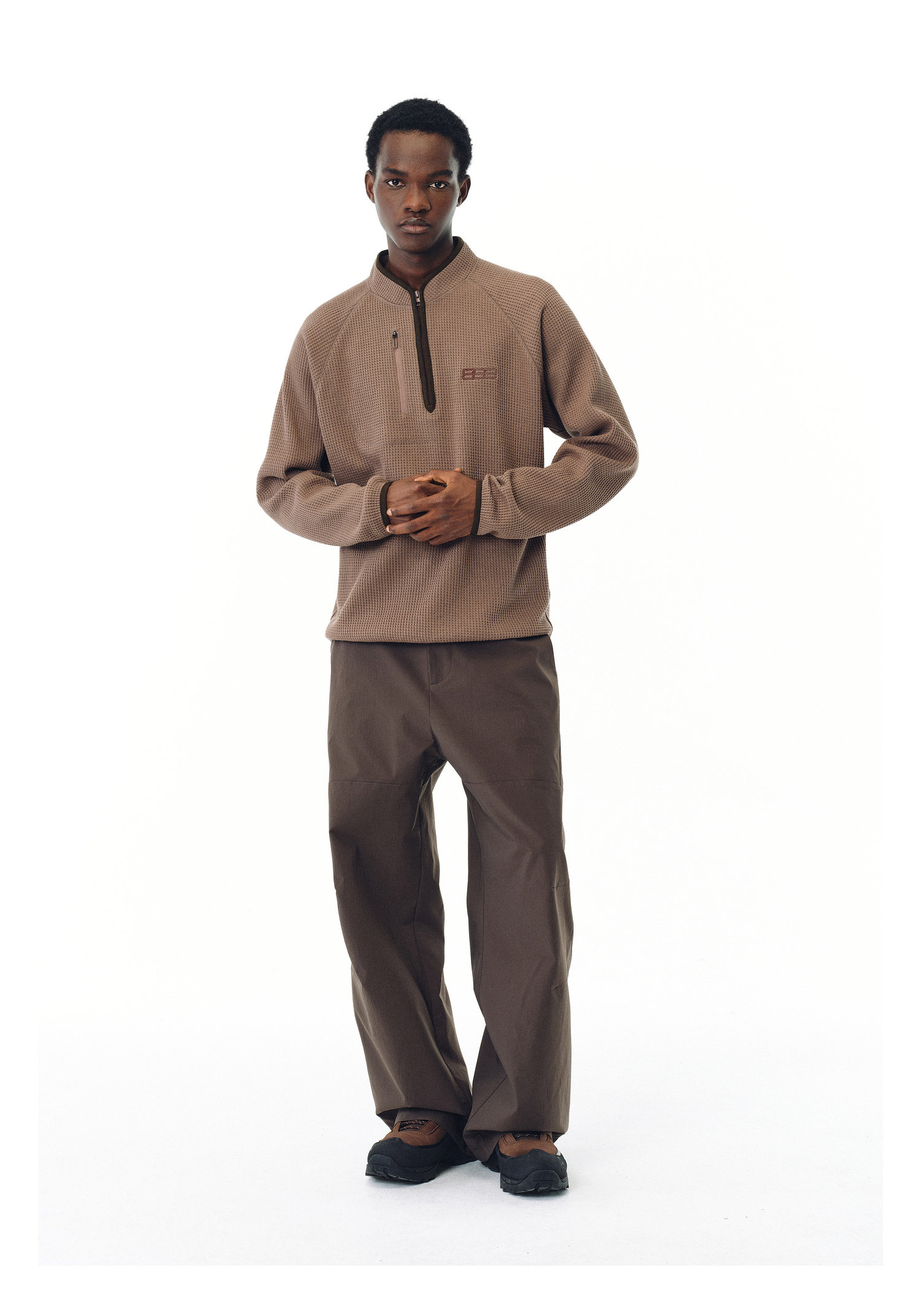 A person stands against a plain white background wearing the EastLyst Heavyweight Magnetic Waffle Knit Pullover Hoodie, matching brown pants, and dark shoes, with hands clasped in front.