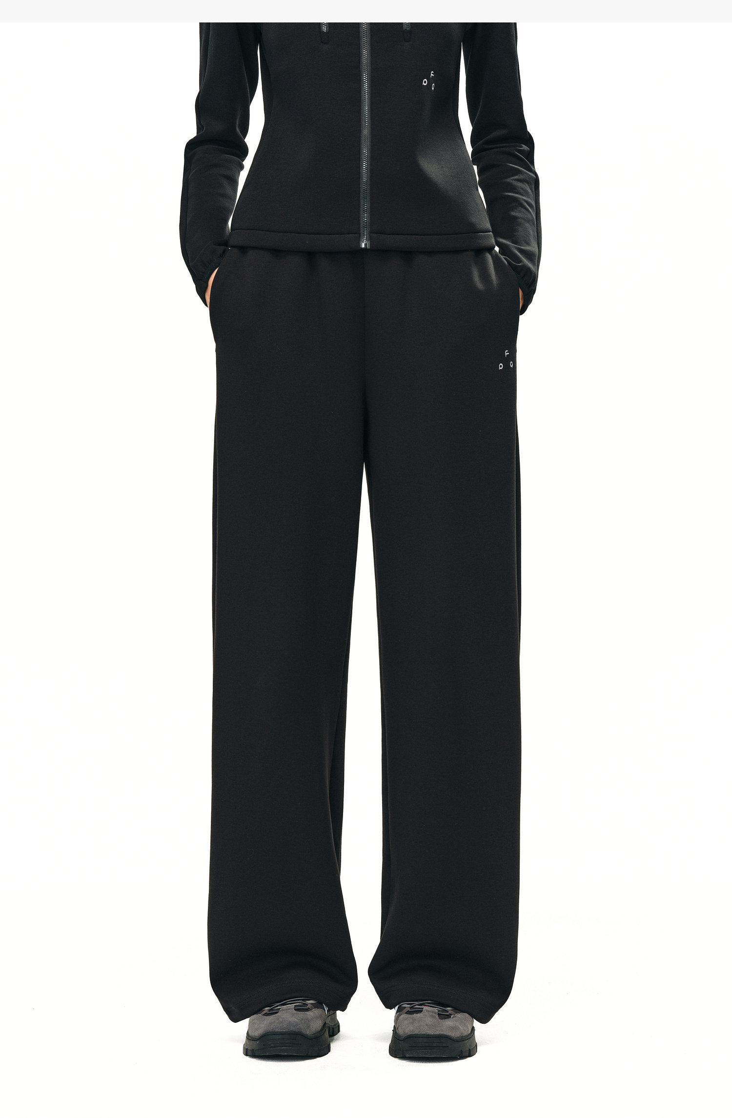 A person stands against a plain white background wearing EastLyst Double-Layer Air-Knit Wide Leg Pants in black with a black zip-up jacket and athletic shoes, hands in pockets. The pants feature a loose, silky silhouette.