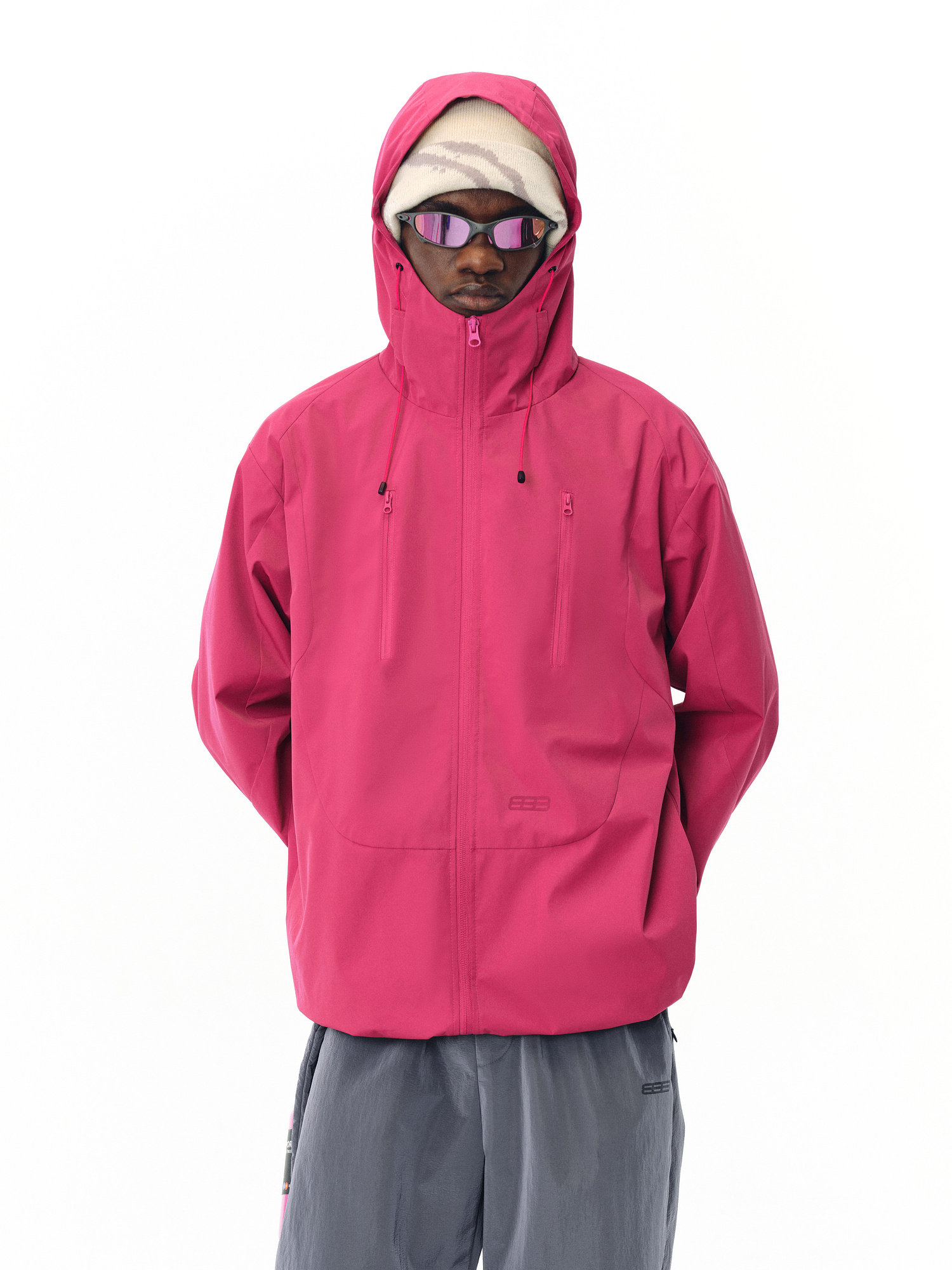 An individual models the EastLyst Best All-Rounder Jacket in bright pink, paired with a light beige hat, sunglasses, and gray pants, standing against a plain white background.