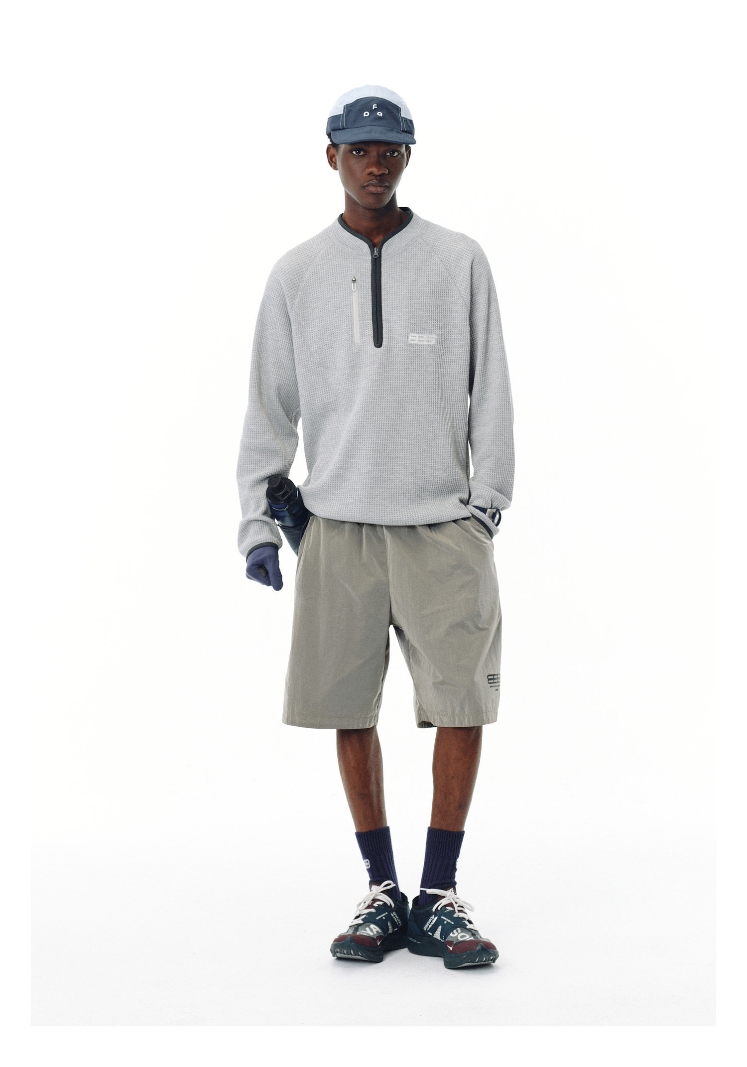 A person in an EastLyst Heavyweight Magnetic Waffle Knit Pullover Hoodie, blue cap, beige shorts, dark navy socks, running shoes, and a single blue glove stands against a plain white background.
