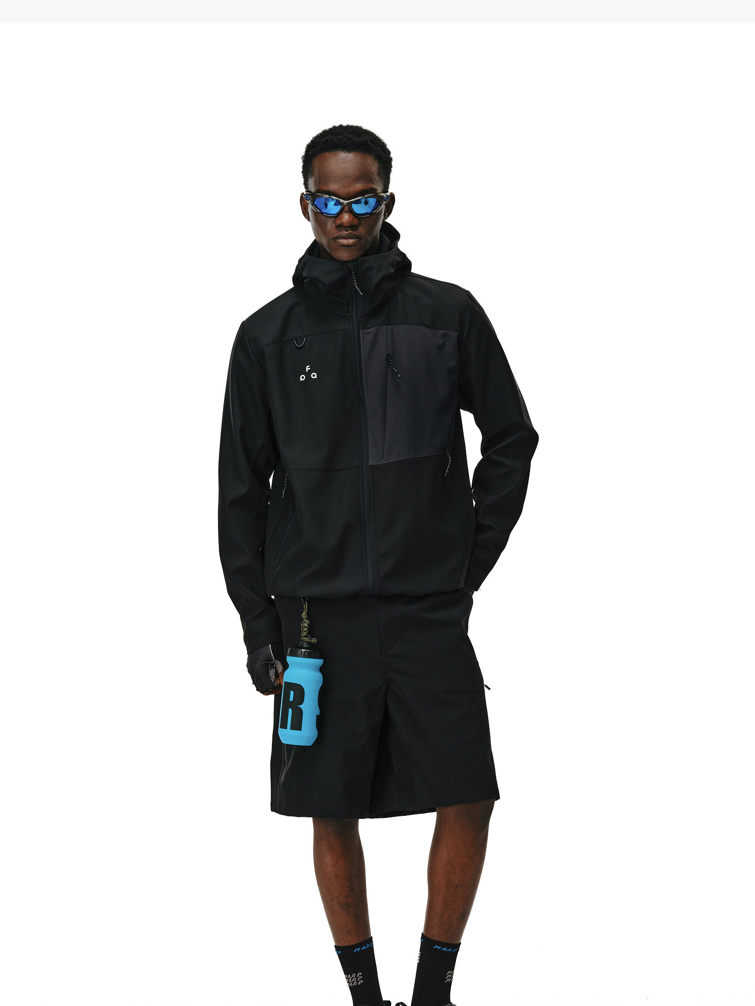 A person wearing the EastLyst Color-Block Water-Resistant Softshell Jacket, black shorts, sunglasses, and gloves stands holding a large blue water bottle with an "R" against a plain white background.
