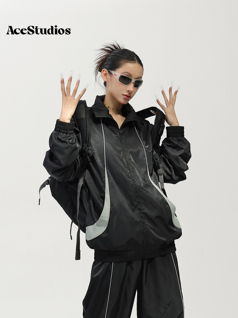 Model wearing a black sporty windbreaker and matching pants with sunglasses and a backpack.