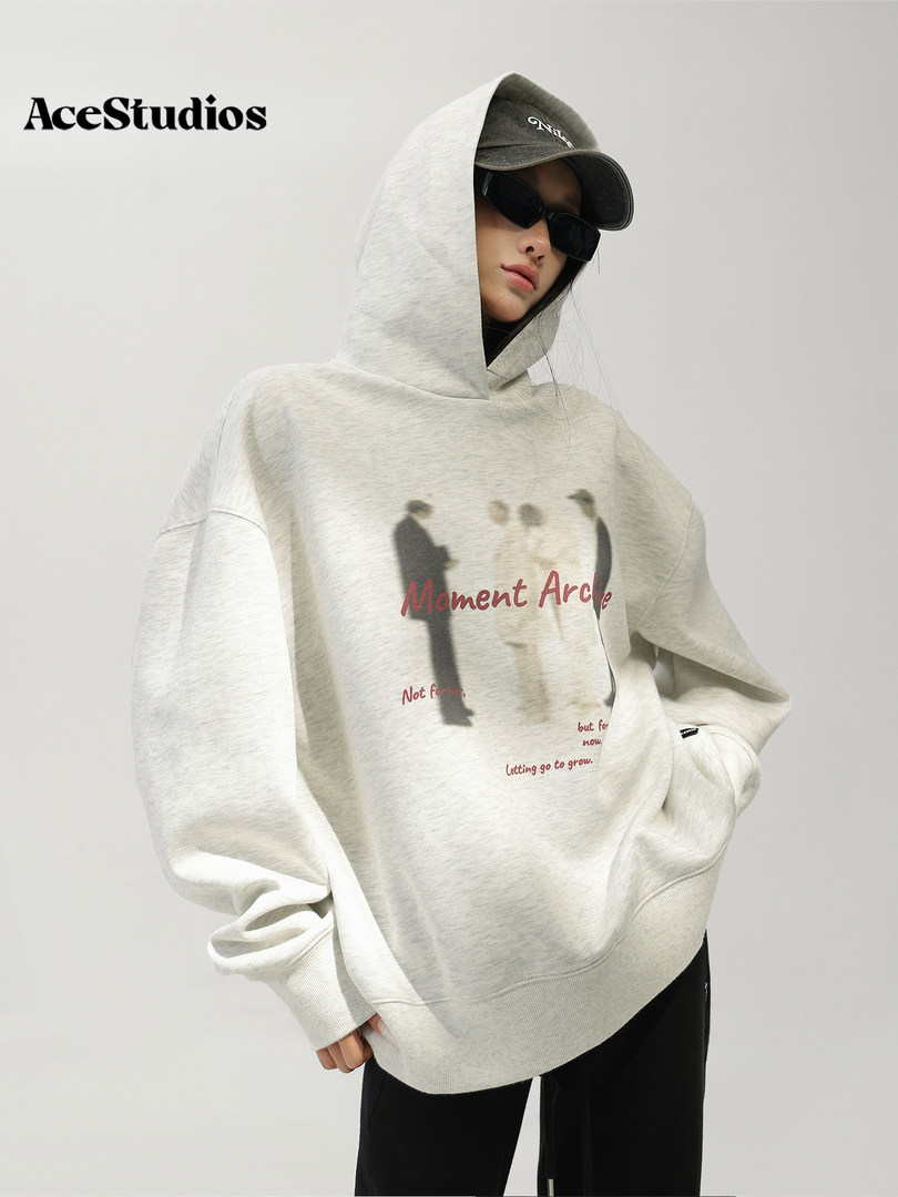 Model wearing an oversized light gray graphic hoodie with hood, cap, and black pants