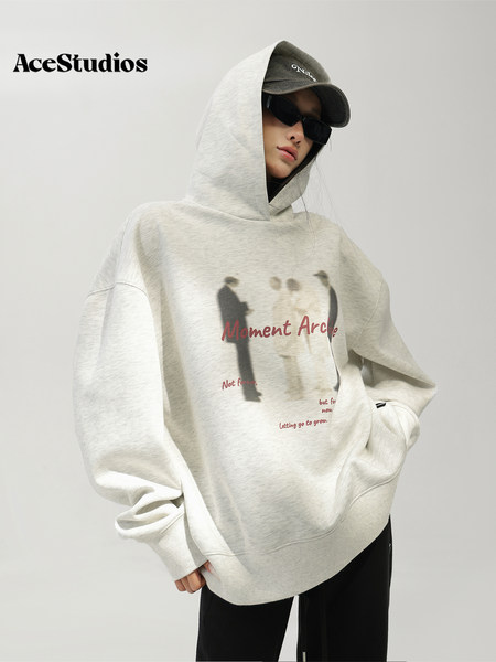 Model wearing an oversized light gray graphic hoodie with hood, cap, and black pants