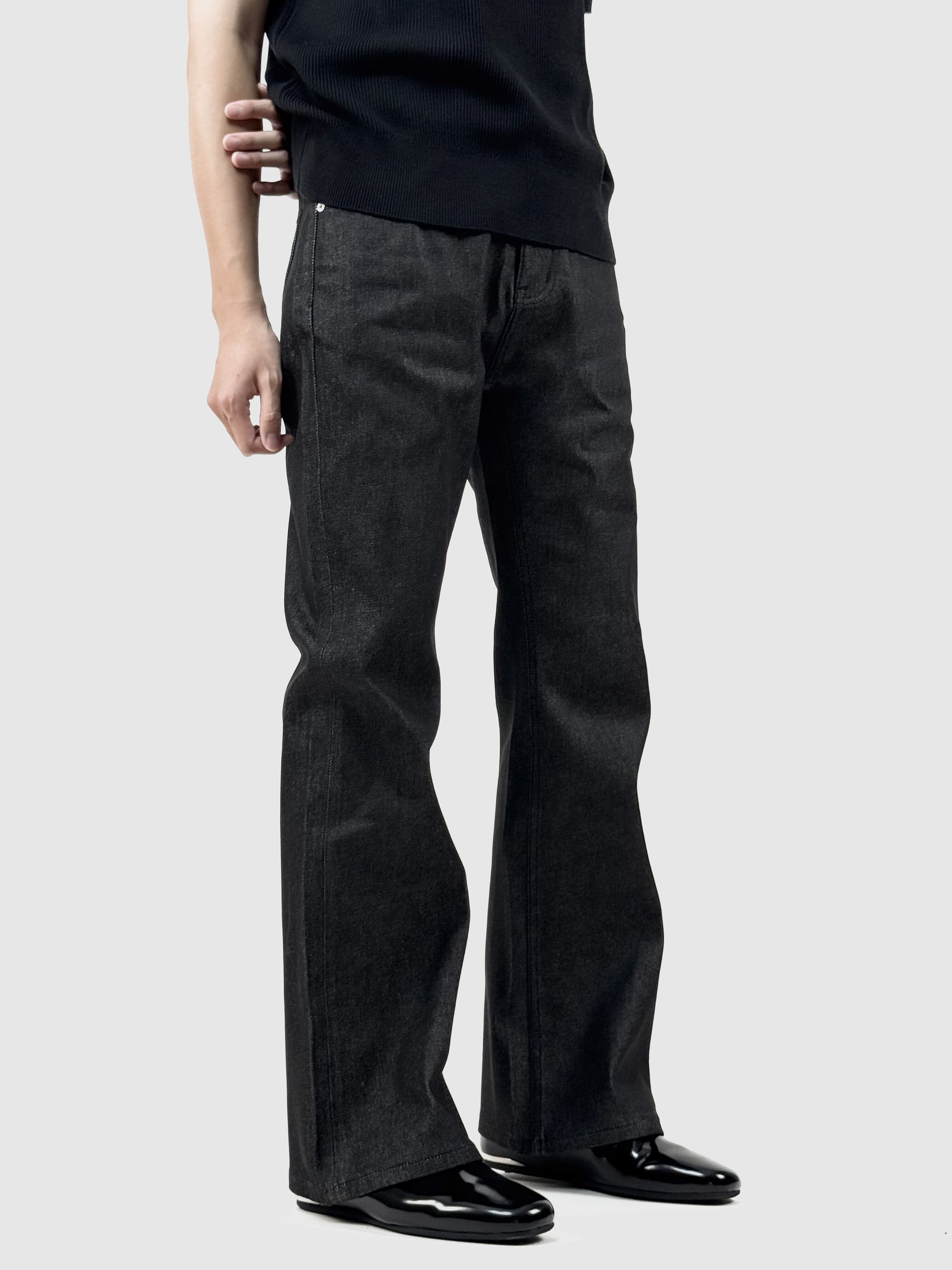 A person models EastLyst’s Colorfast Black Flared Jeans with contrast stitching, paired with a black top and shiny black shoes, standing against a light gray background. The photo is cropped at the shoulders for anonymity.